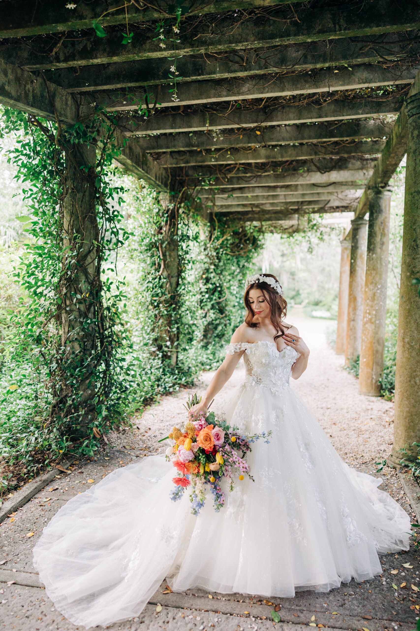 A bride in a long flowing dress smiles down her shoulder to her vibrant bouquet while walking under a vine covered walkway at her Airlie Gardens wedding