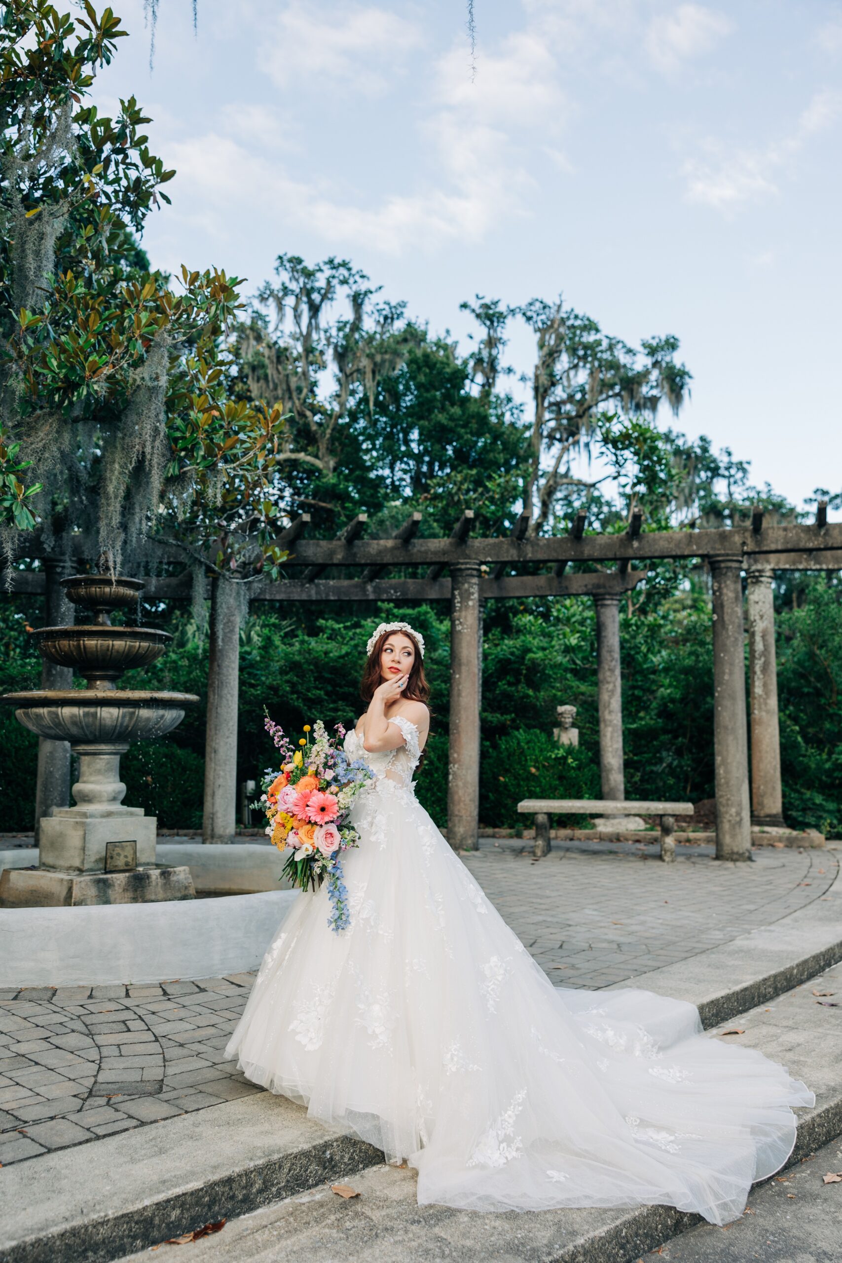 A bride touches her face while standing by a garden fountain