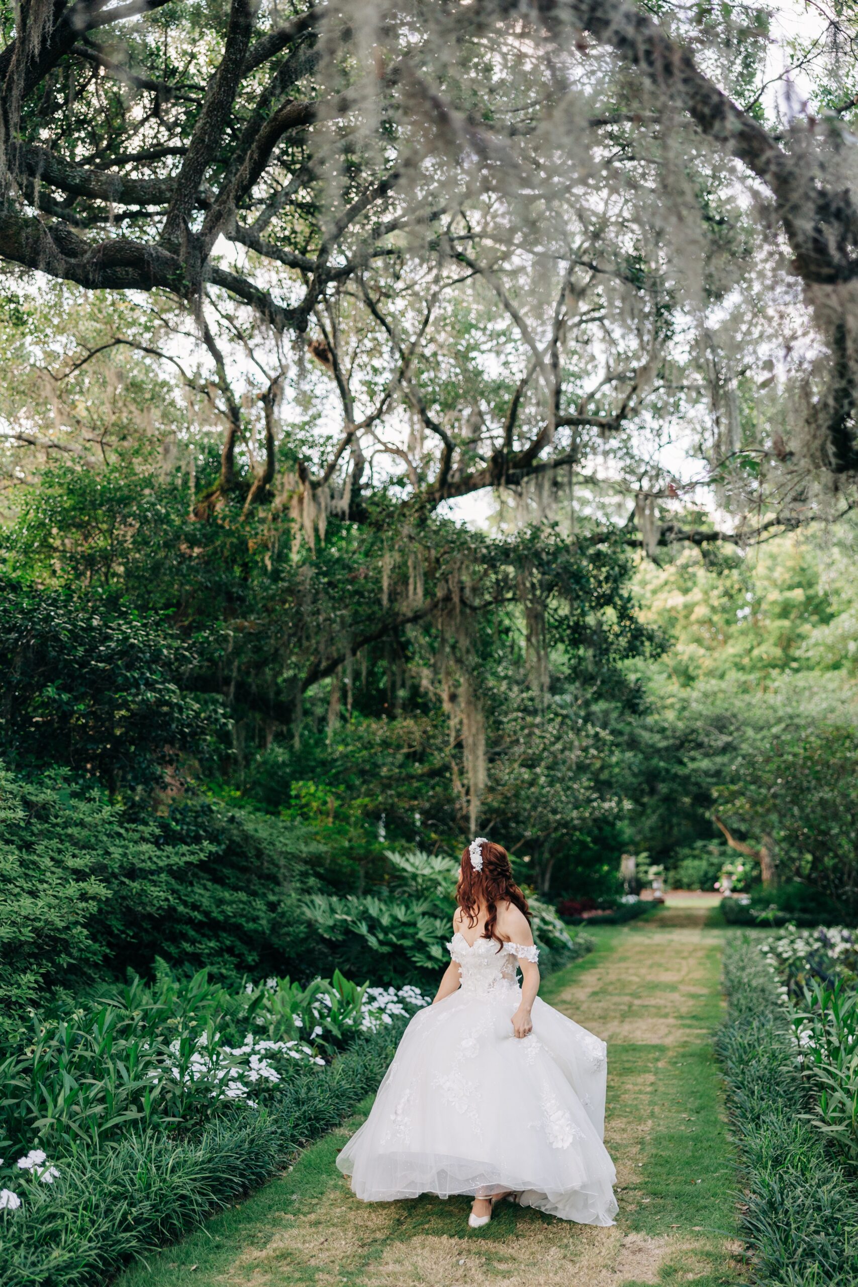 A bride looks over her shoulder while holding her train and walking through a path at her Airlie Gardens wedding