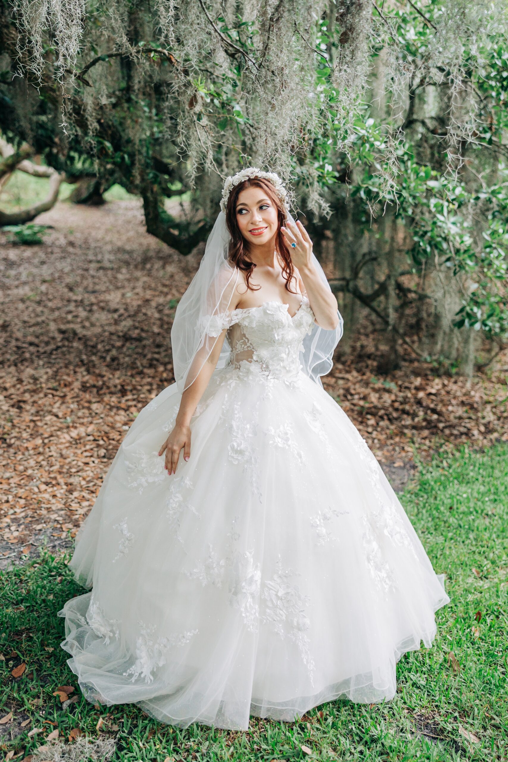 A bride shows off her ring while walking in her dress under an oak tree