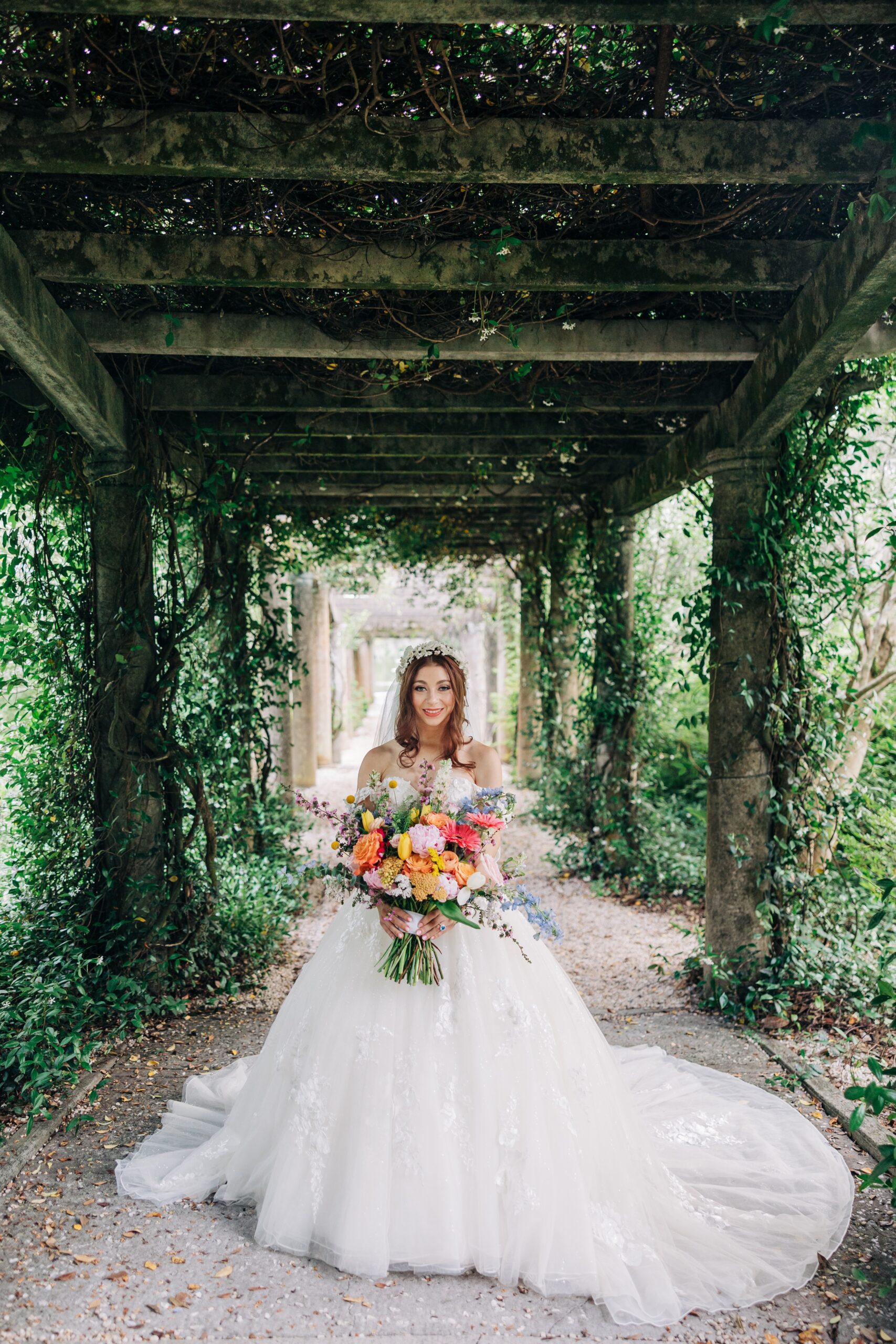 A bride smiles while holding her colorful bouquet in a vine covered walkway at an Airlie Gardens wedding