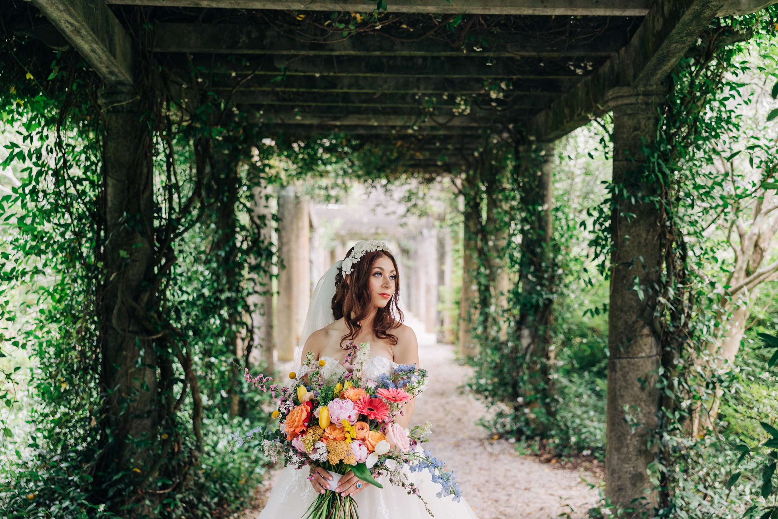 A bride gazes over her shoulder while walking under a vine covered walkway at her Airlie Gardens wedding