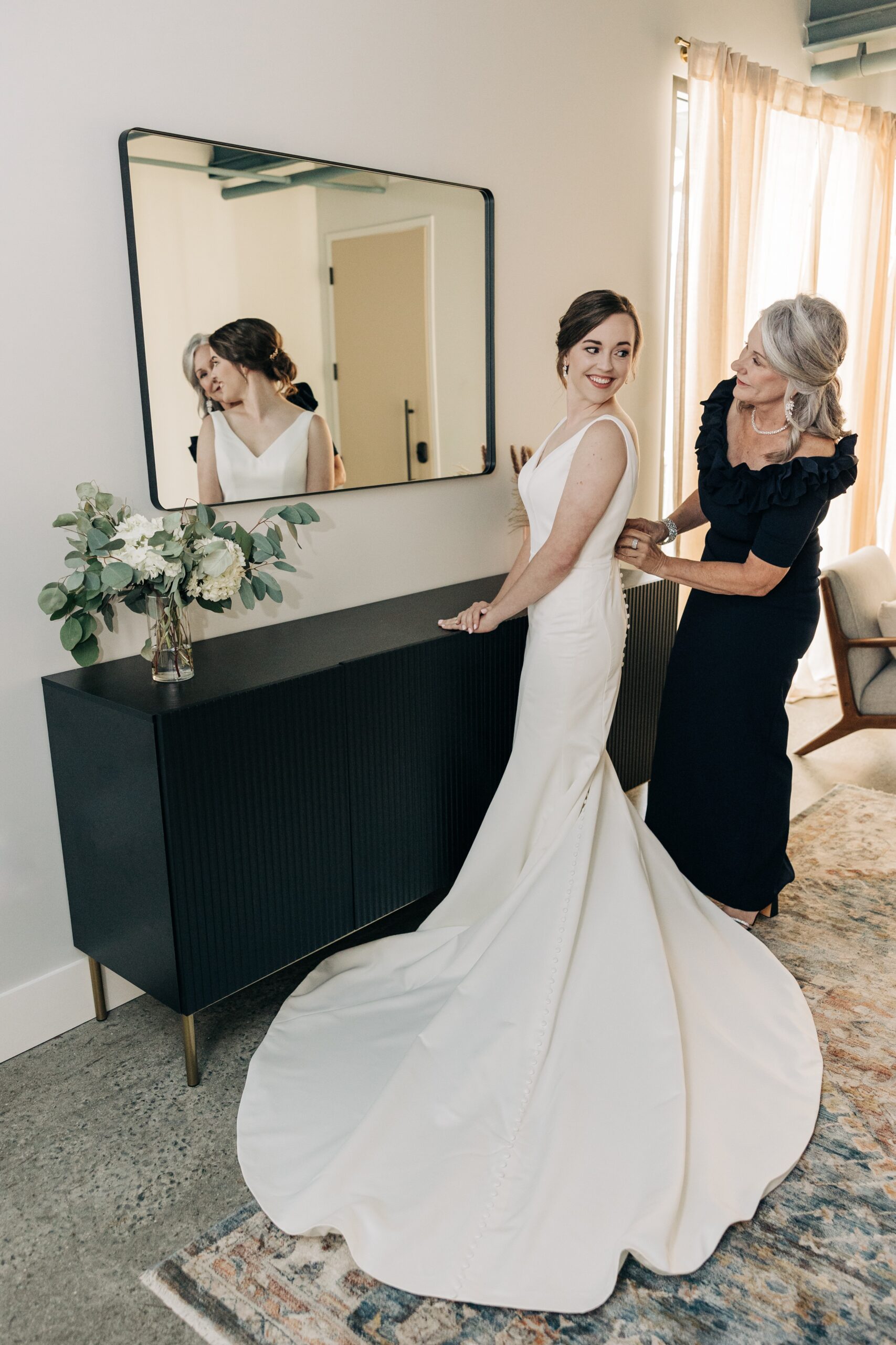 A bride gets help from mom buttoning up her dress while standing in a mirror