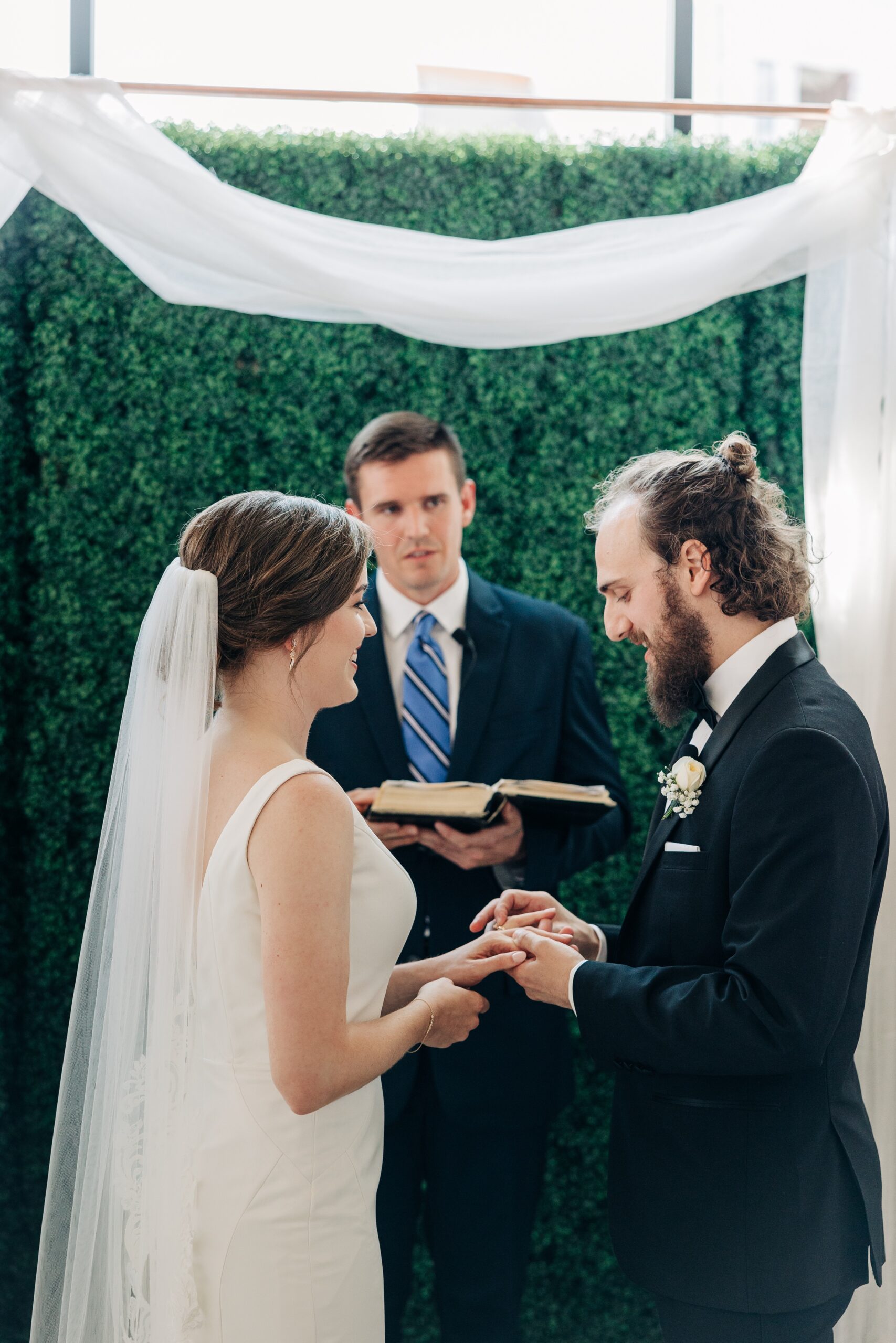 A groom in a black suit puts the ring on his bride's finger during their pine wedding venue ceremony