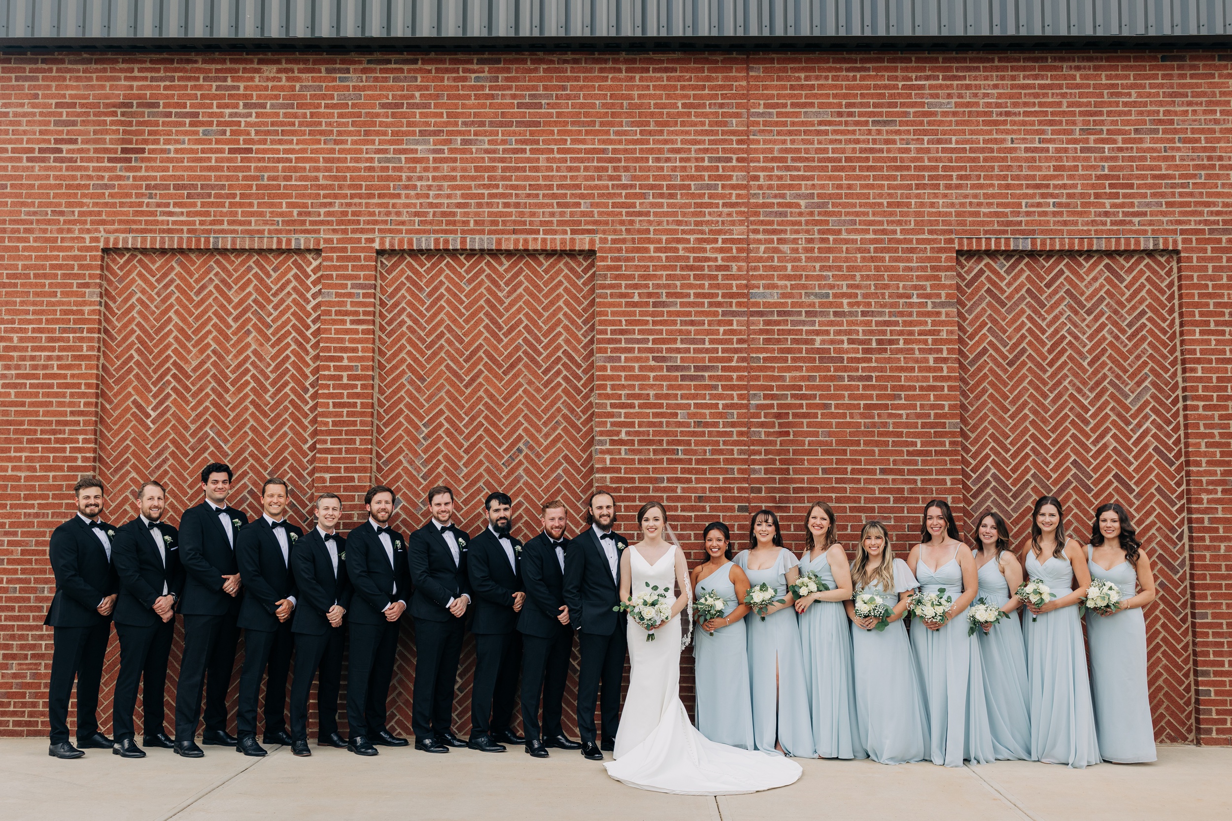 Newlyweds stand with their large wedding party against a brick wall at the pine wedding venue