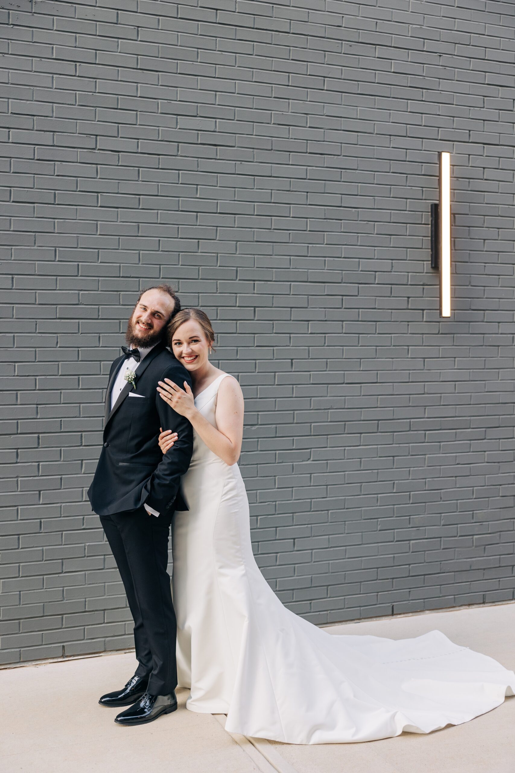 A bride leans on the shoulder of her groom while standing in an alley with a grey brick wall