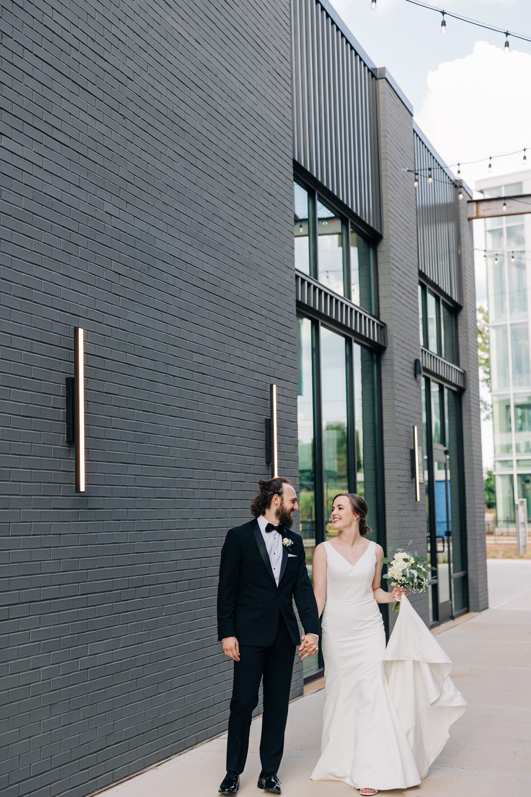 A bride and groom walk hand in hand outside the pine wedding venue while smiling at each other under market lights