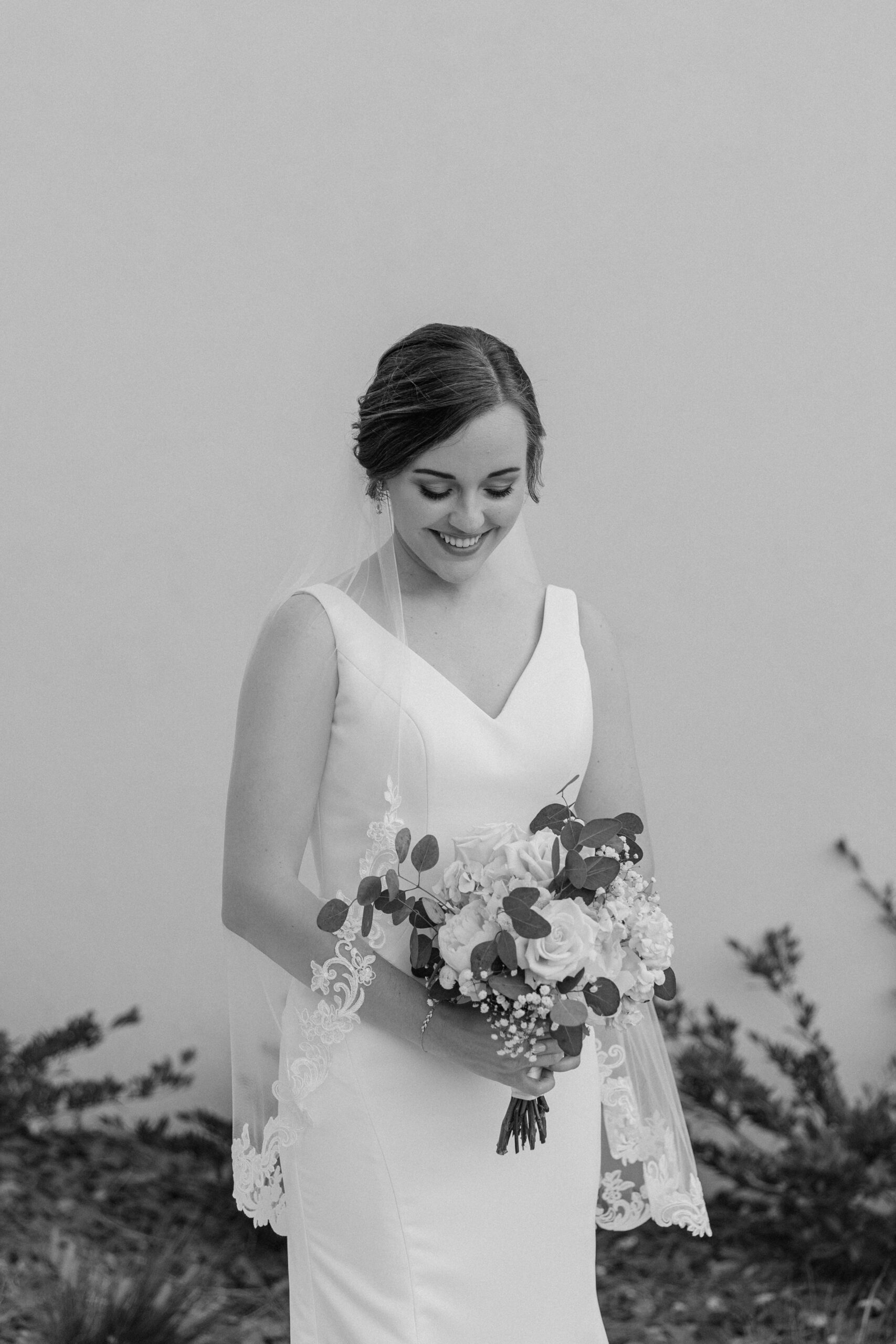 A bride smiles down to her bouquet in black and white while standing outside