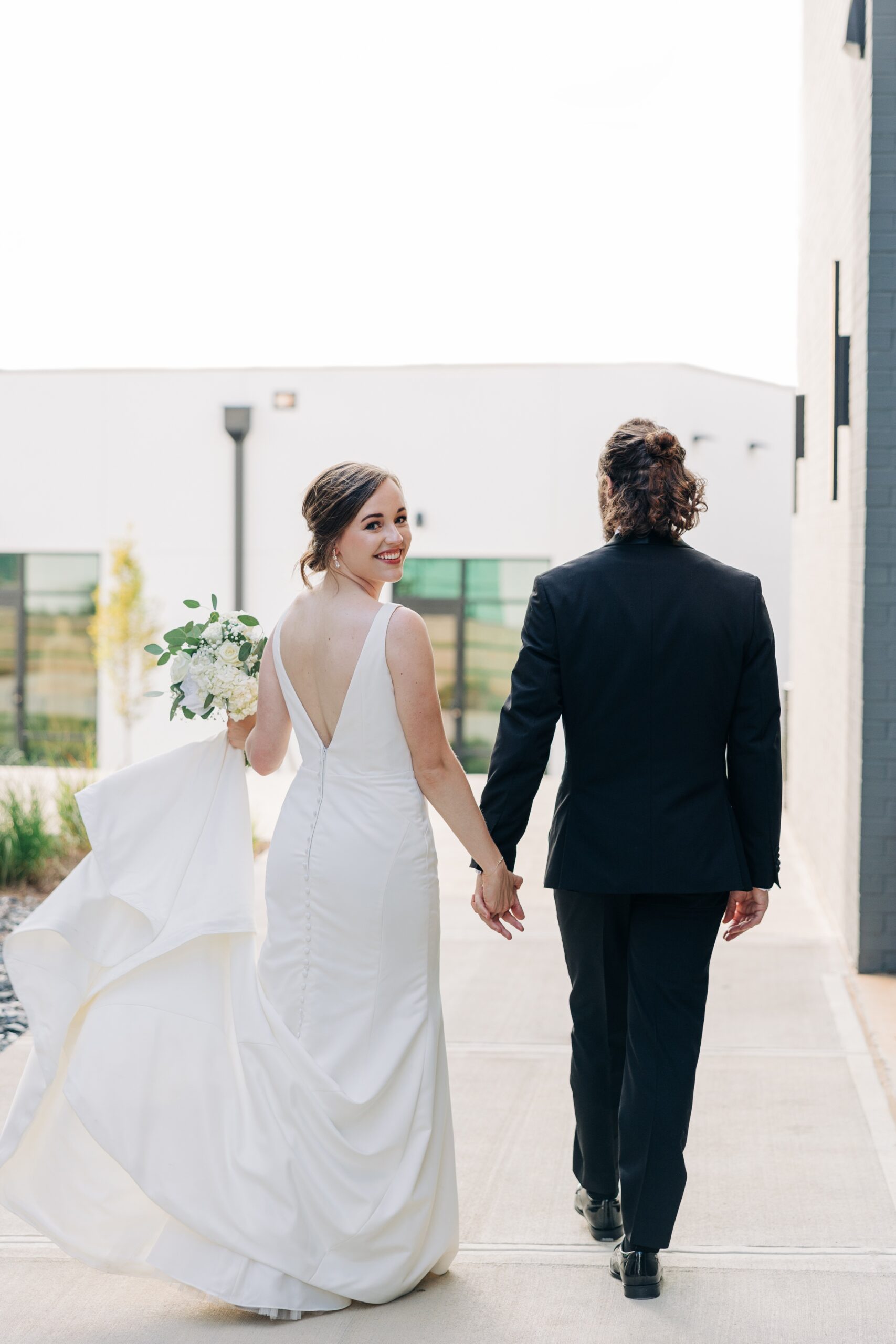 A bride smiles over her shoulder while holding hands and walking with her groom outside the pine wedding venue