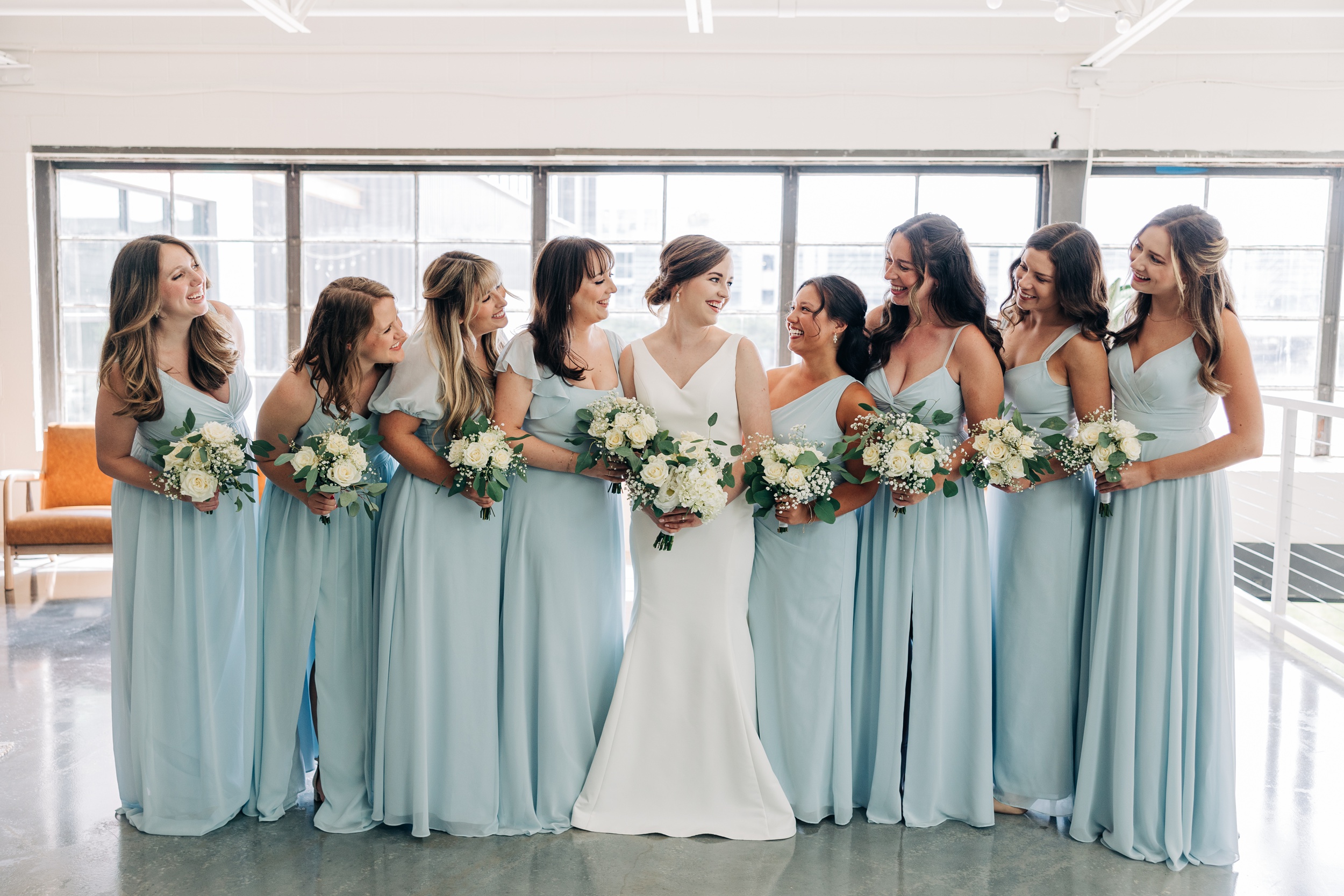 A bride laughs with her many bridesmaids in blue dresses holding their bouquets at the pine wedding venue