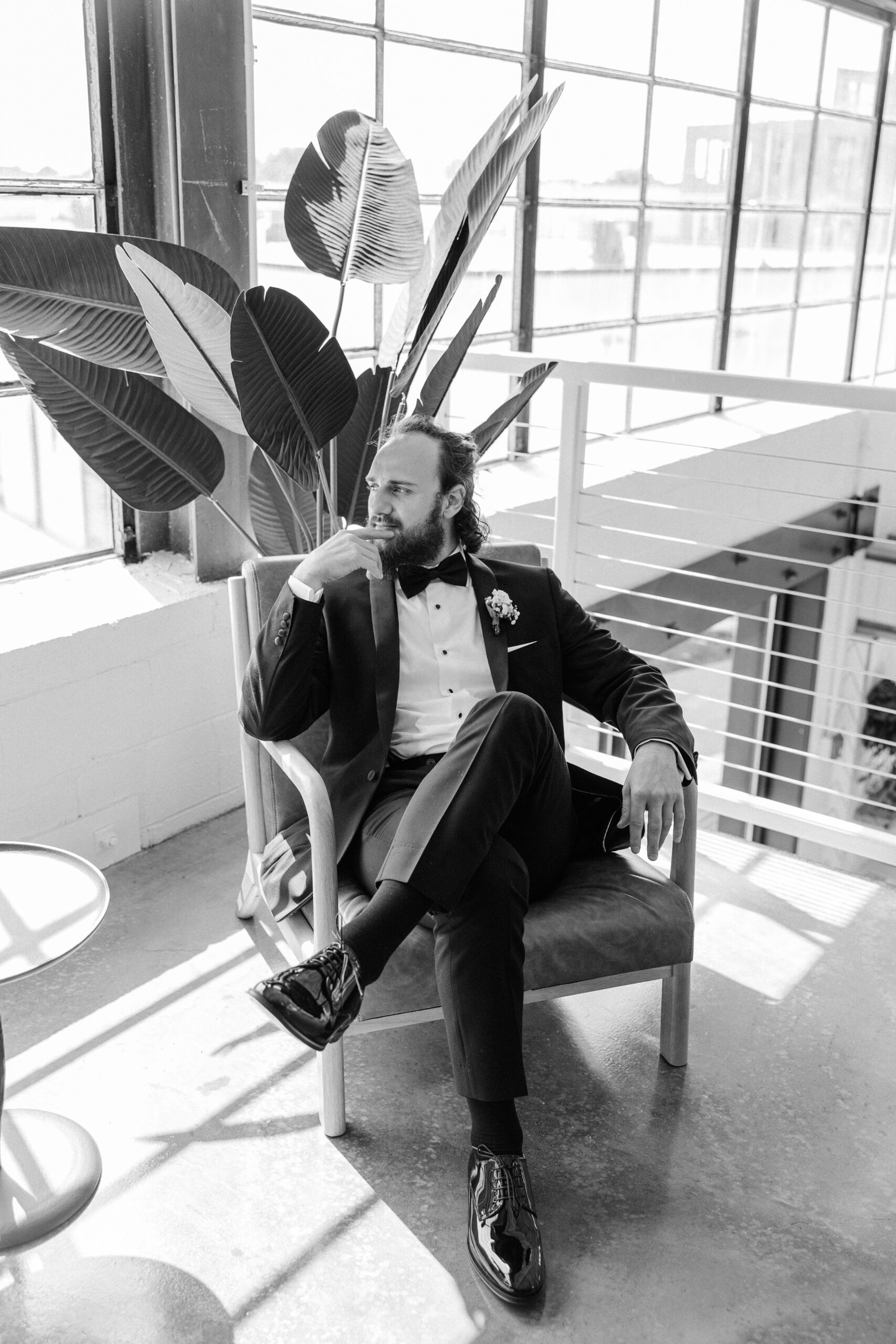 A groom in a black suit sits in a chair gazing out a window on a balcony