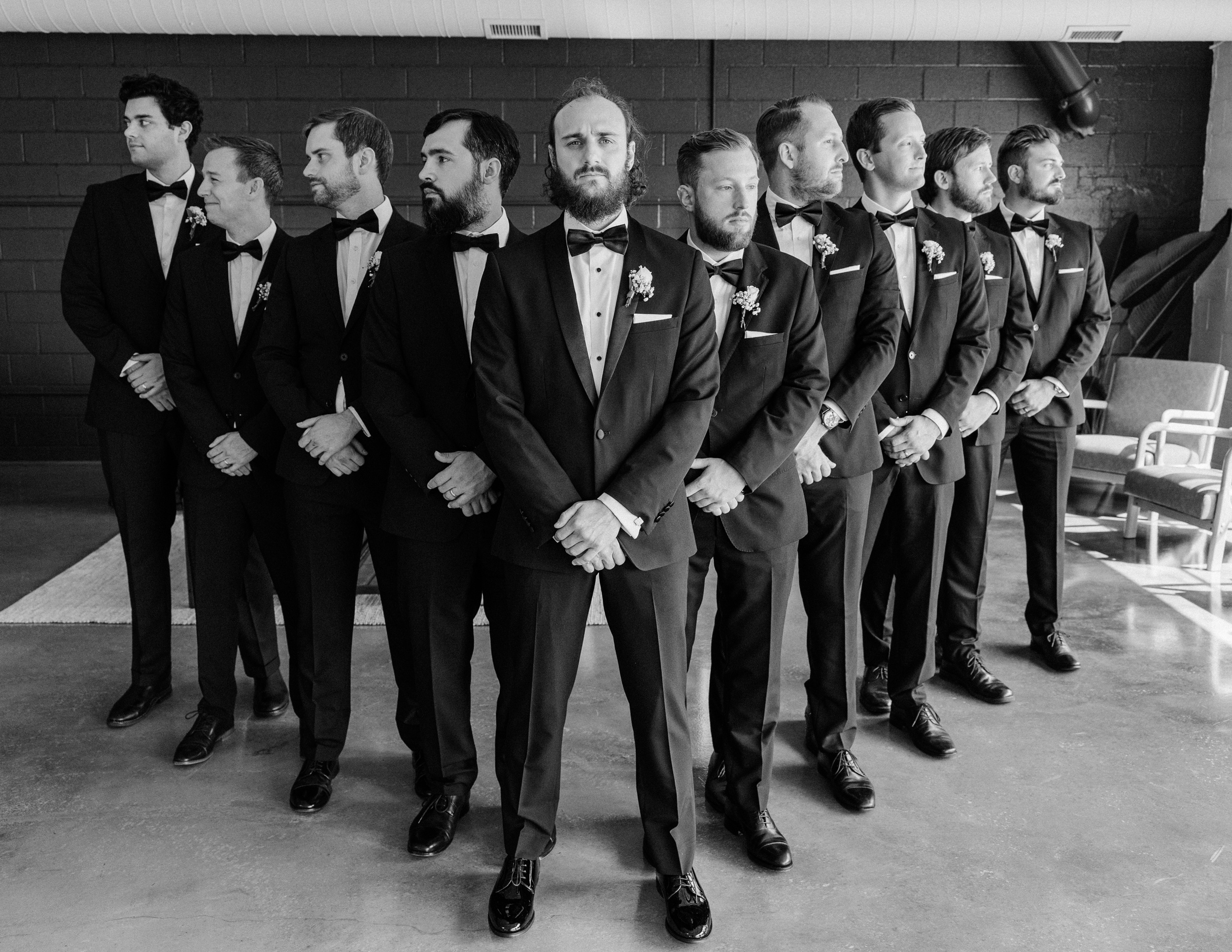 A groom stands with hands crossed with his groomsmen in a V behind him in black and white