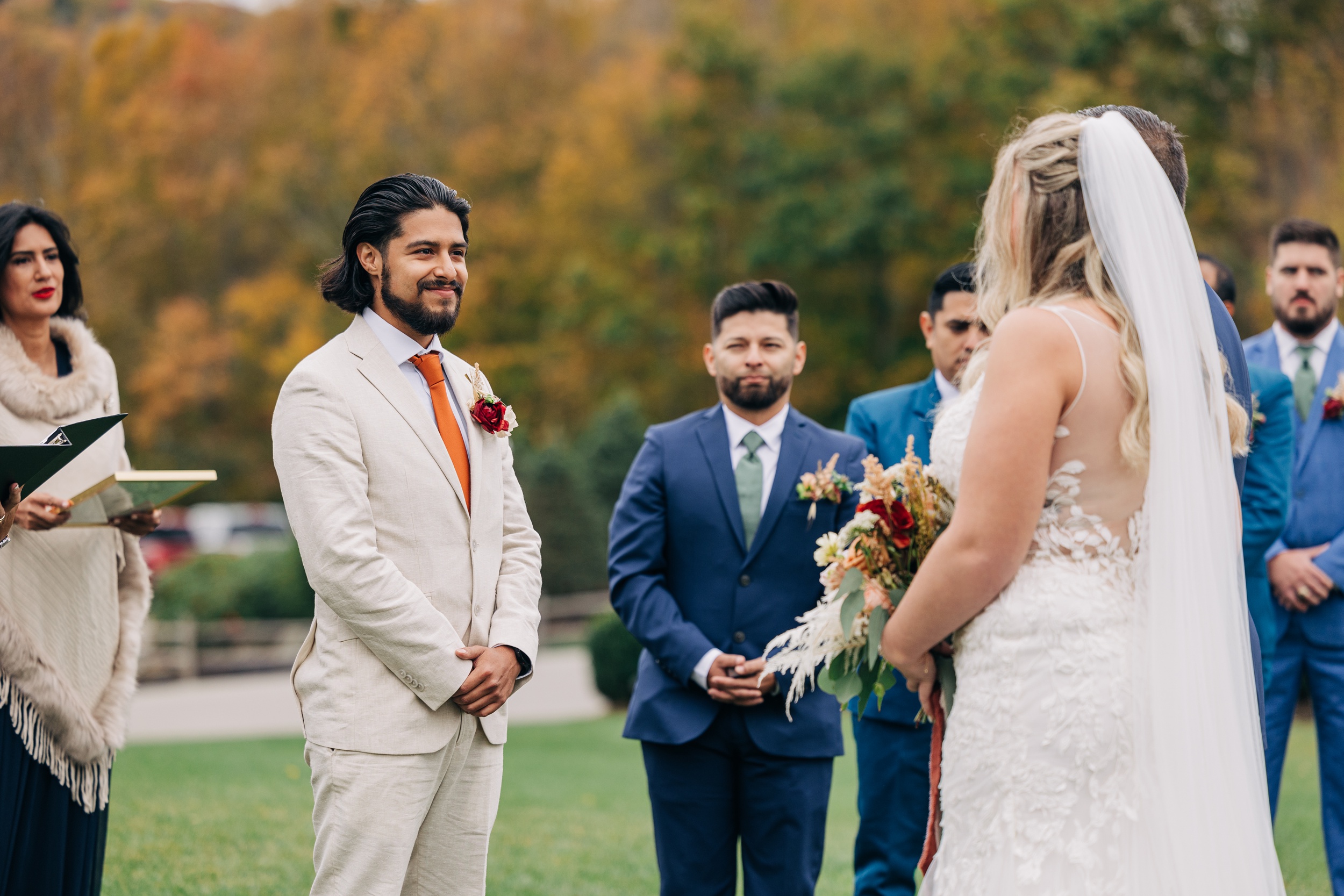 A groom in a tan suit smiles big while seeing his bride approach for their ceremony