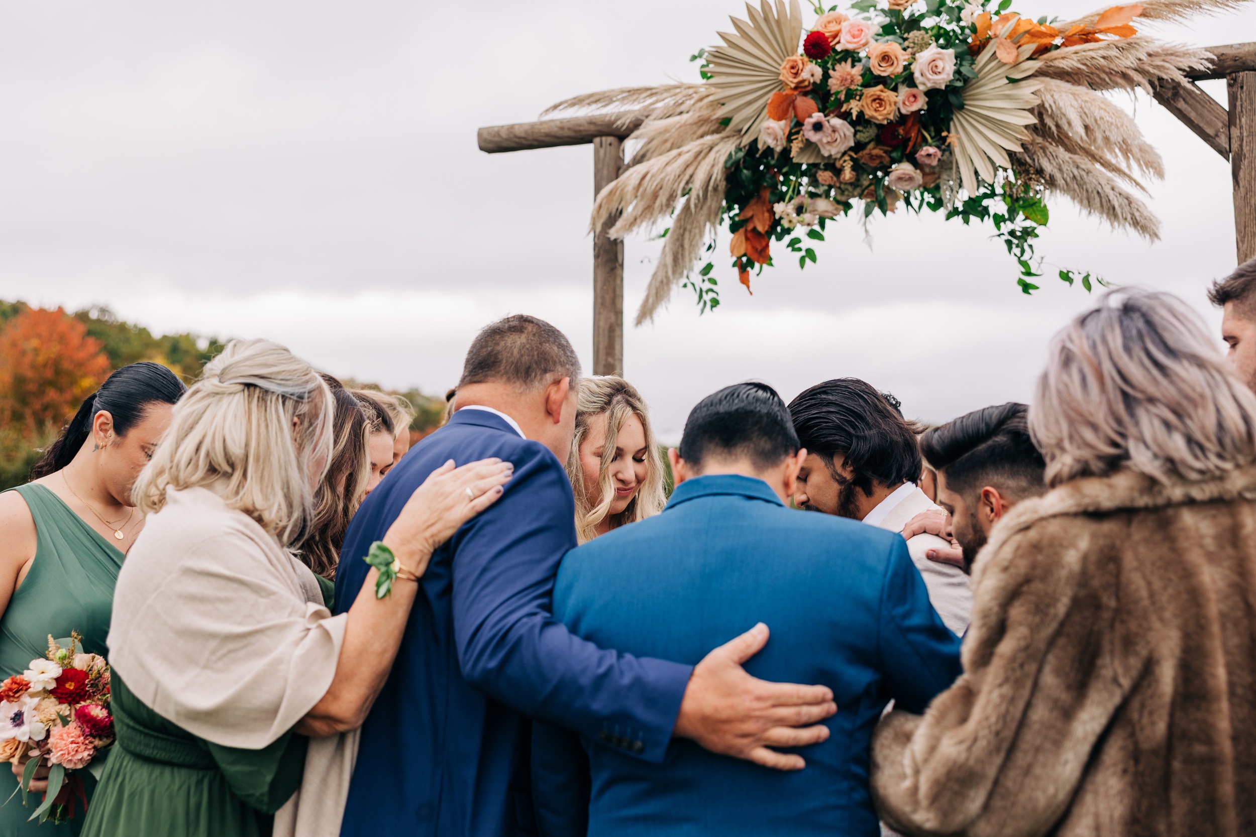 A bride and groom are surrounded by their guests under the wooden arbor for a prayer