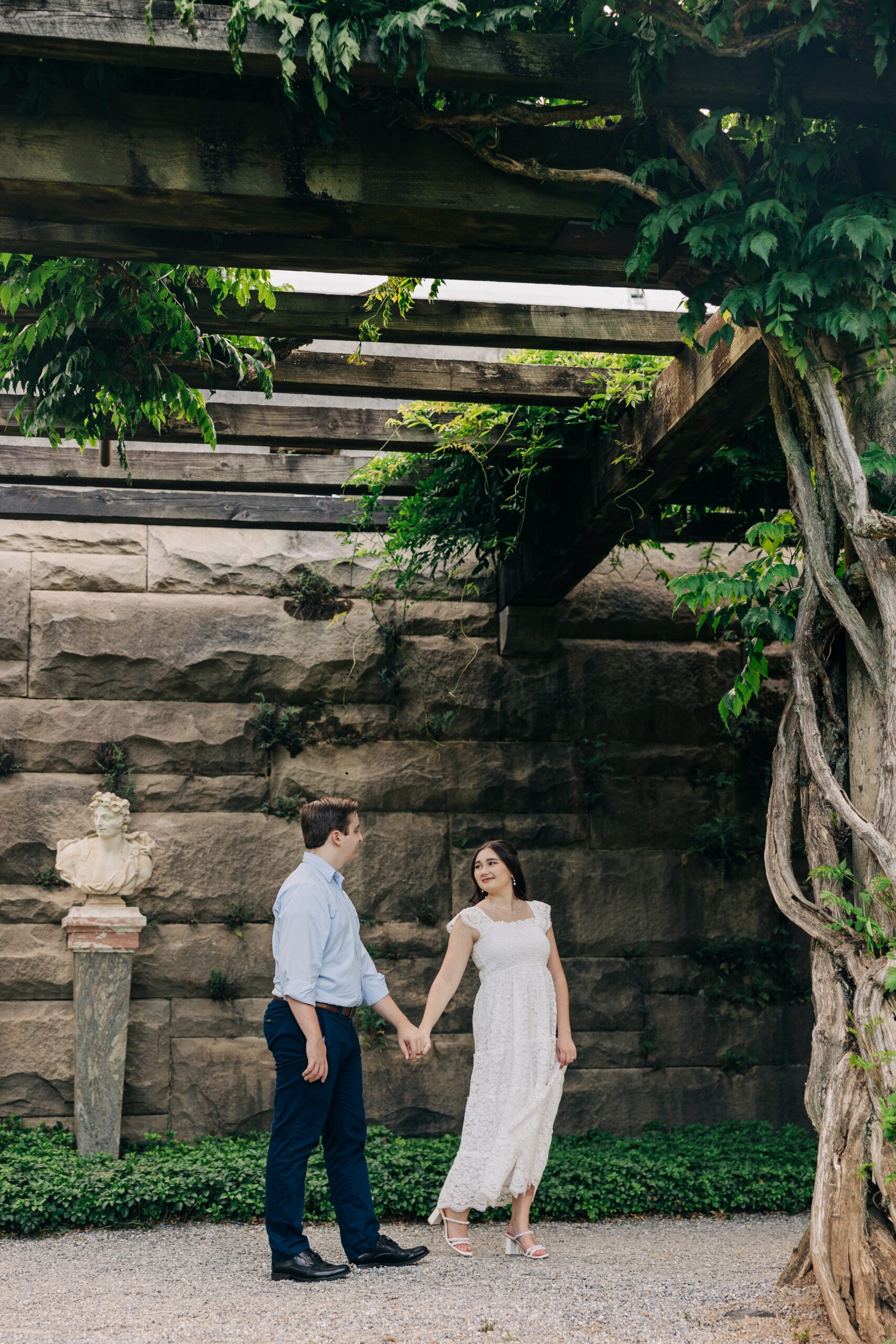 A bride leads her groom by the hand under a garden overhang while smiling back at him