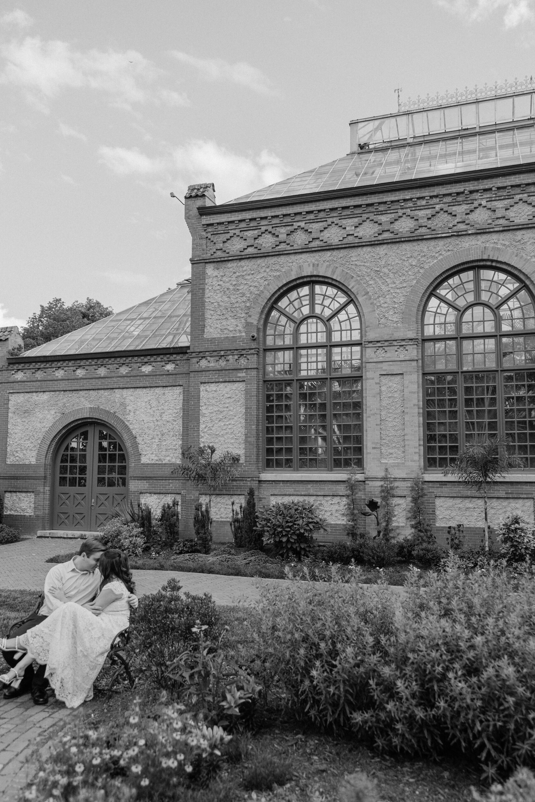 Newlyweds kiss while sitting on a garden bench by an ornate greenhouse