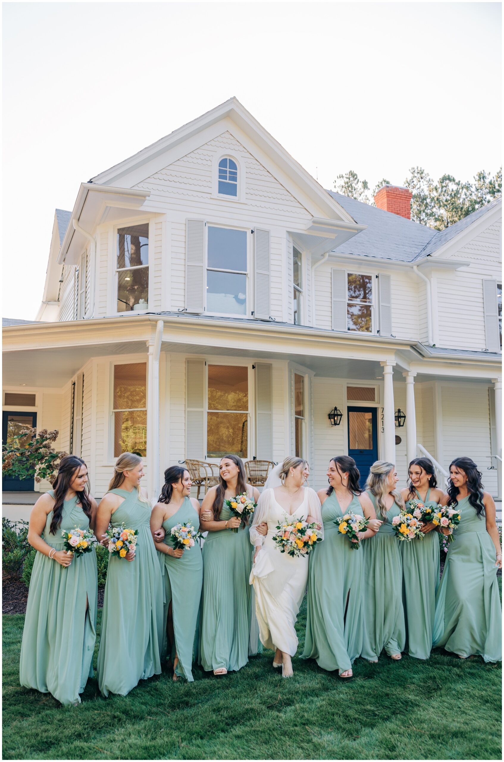 A bride walks with her bridal party in green dresses in the lush lawn of the upchurch wedding venue