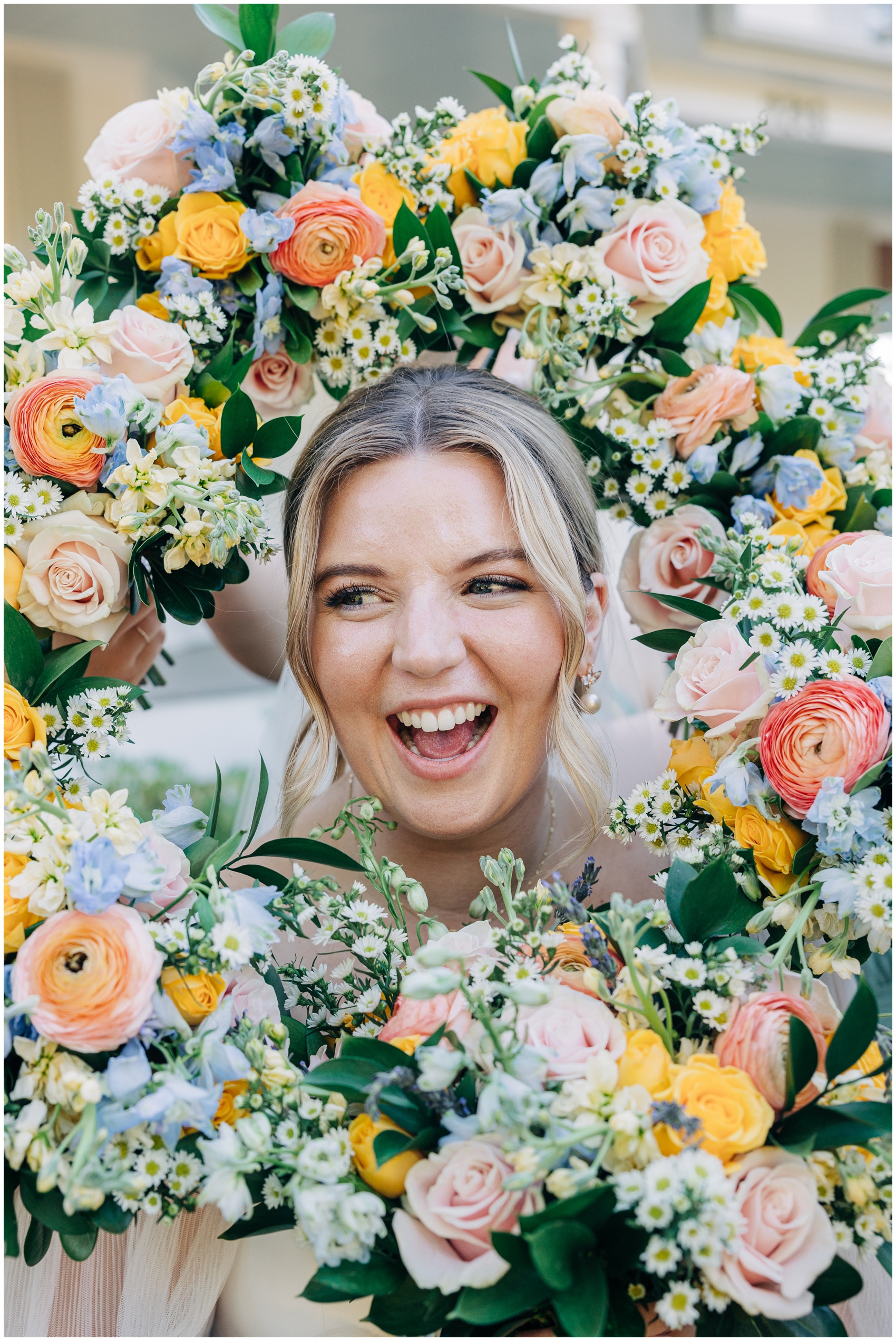 A bride laughs big while looking through a colorful floral wreath