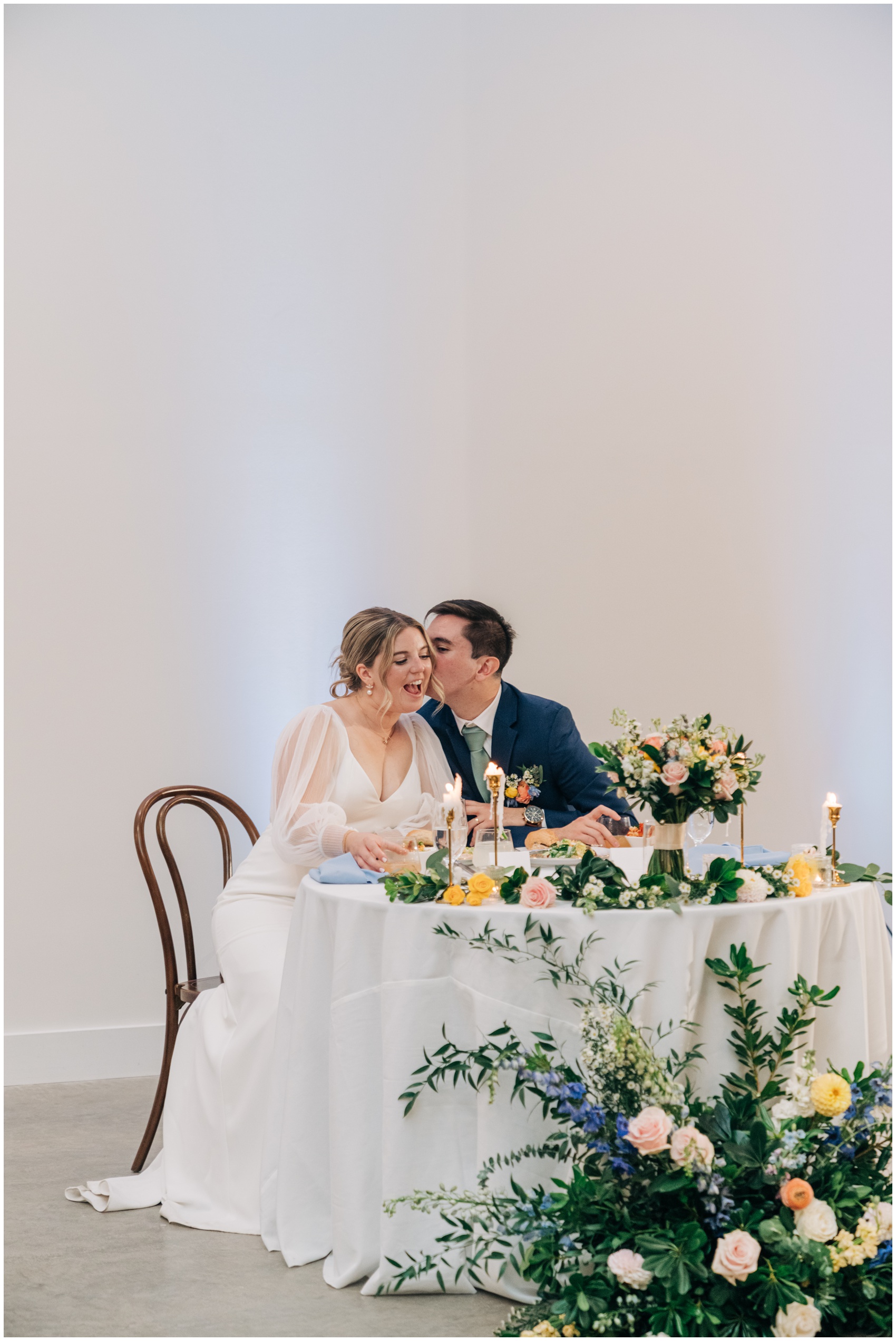 A groom kisses the cheek of his bride as they sit at their head table covered in flowers