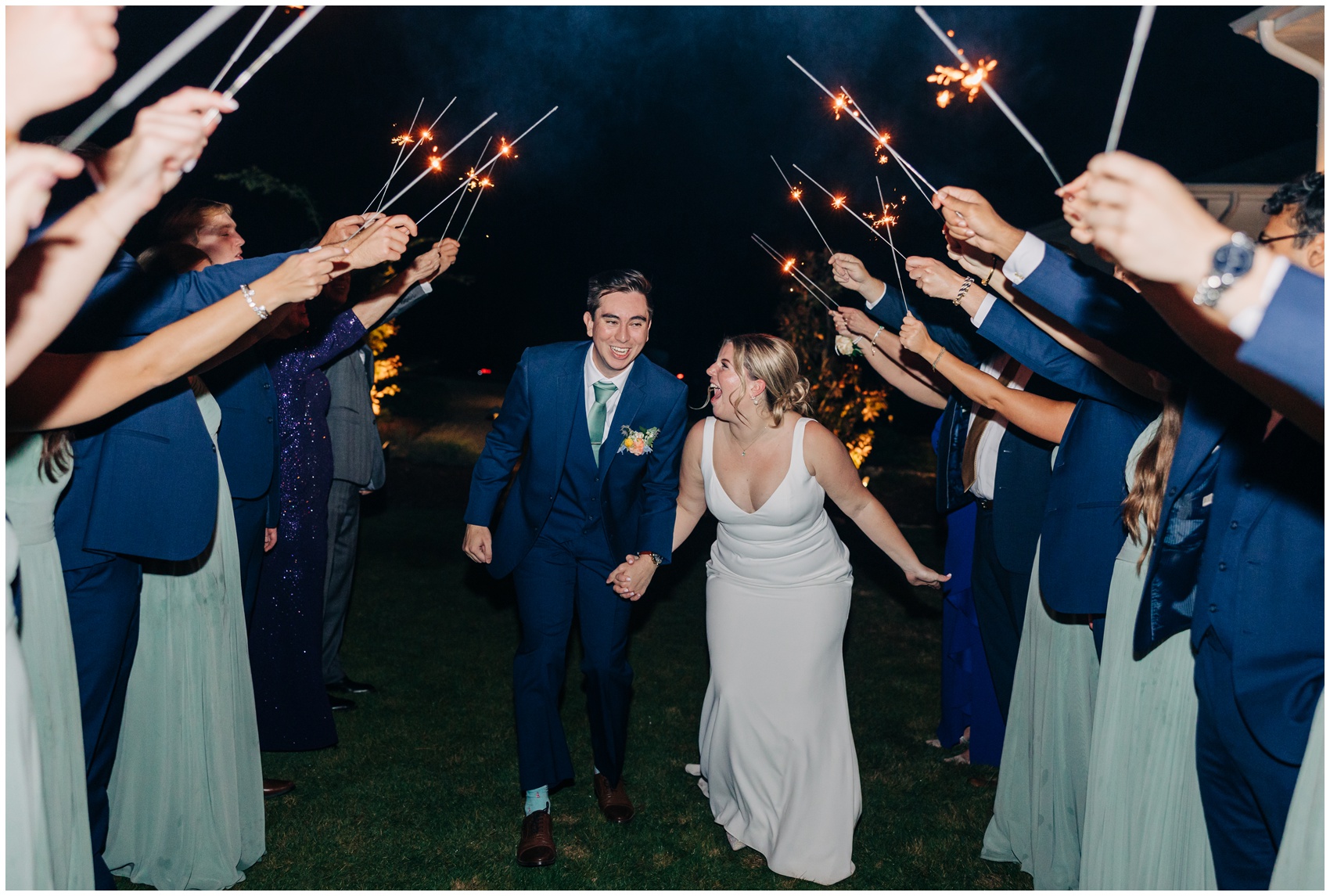 A bride and groom laugh big while holding hands and walking under an aisle of sparklers to exit their wedding