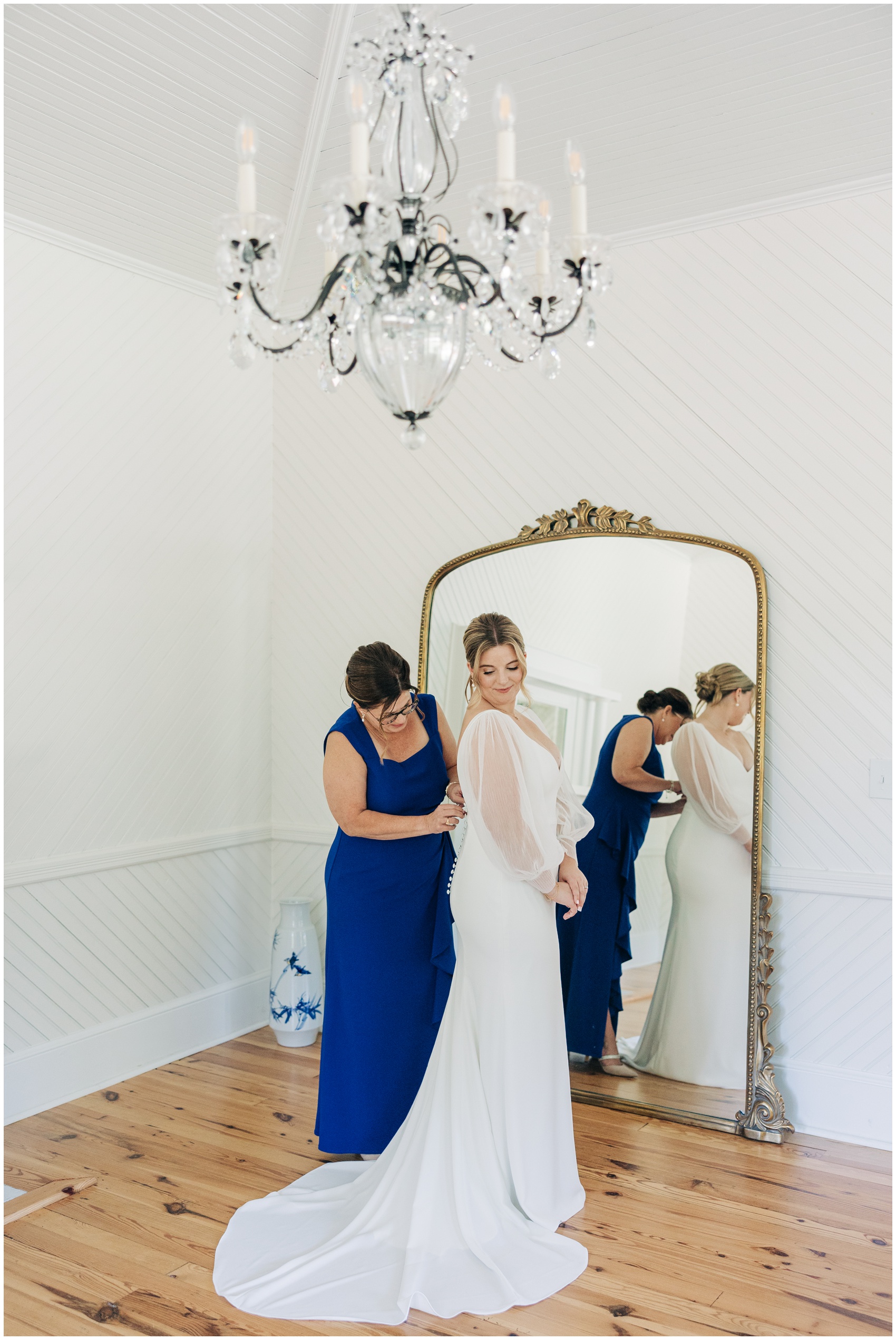 Mom helps button in the bride in a large ornate mirror under a chandelier