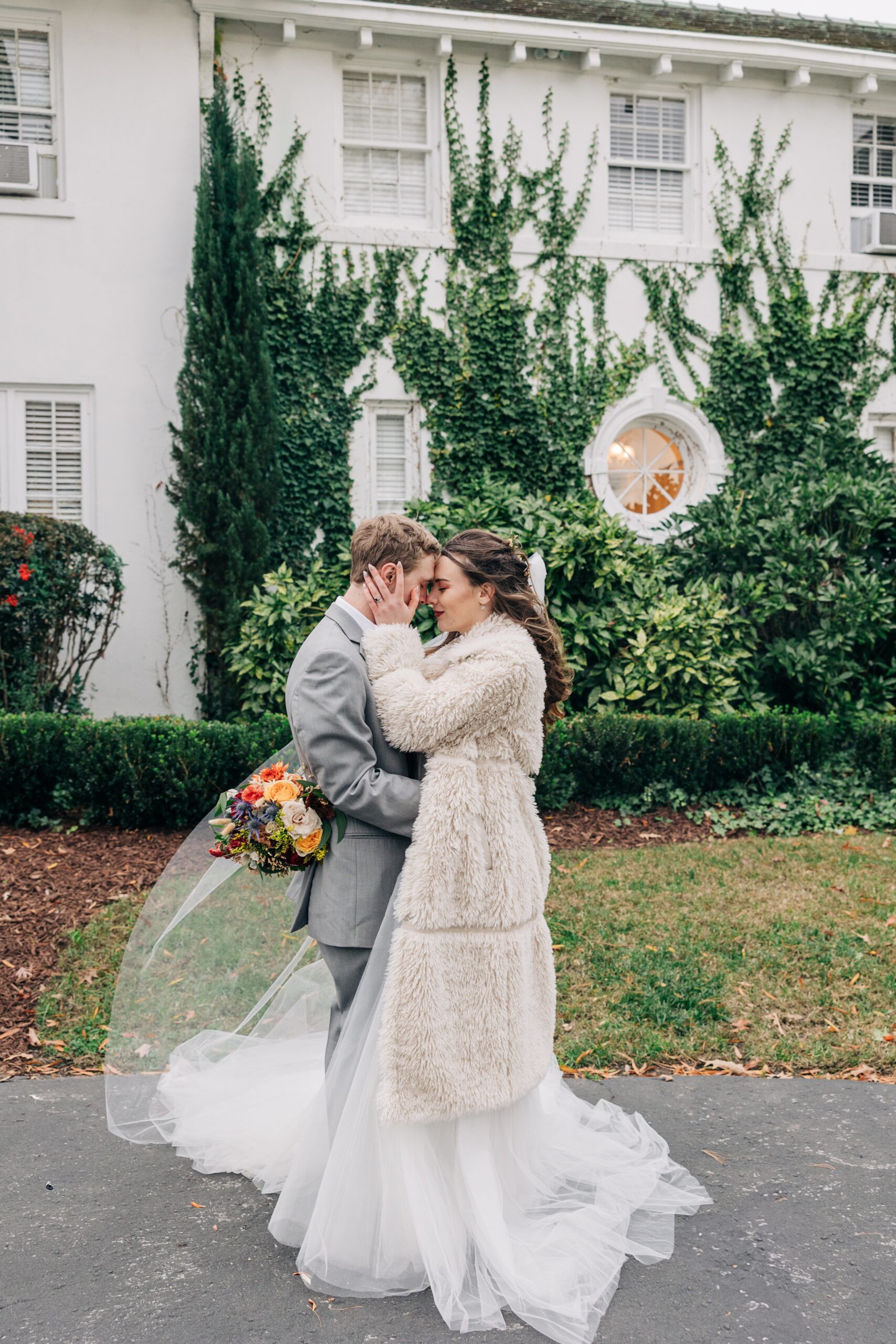 A happy couple snuggle in a fur coat and grey suit in the gardens of The Finch House