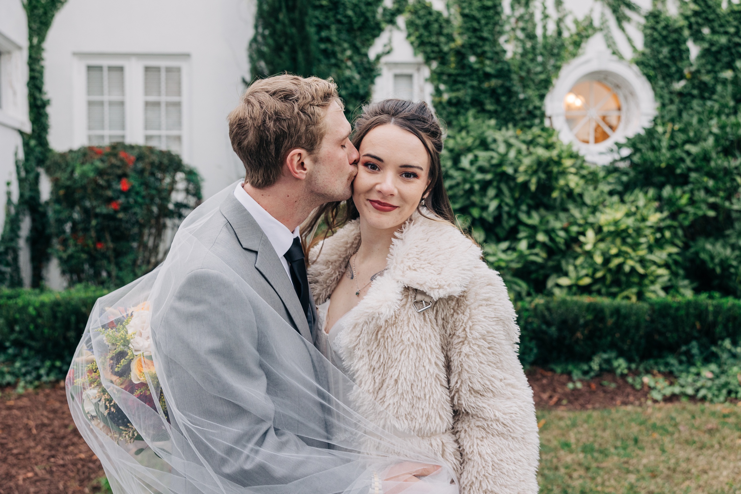 A bride smiles while her groom kisses her cheek in a garden