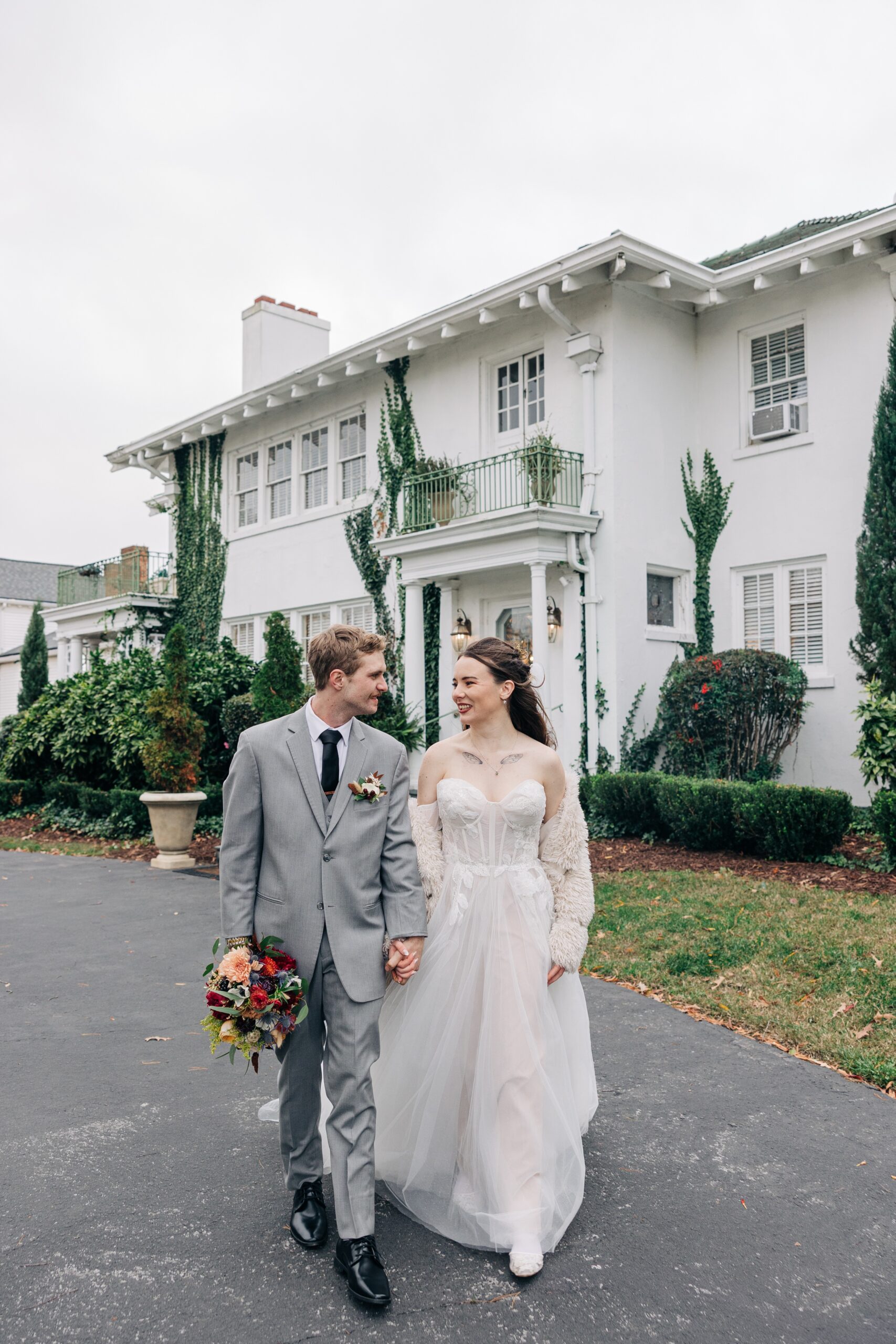 Happy newlyweds walk in the driveway of their wedding venue while smiling and holding hands