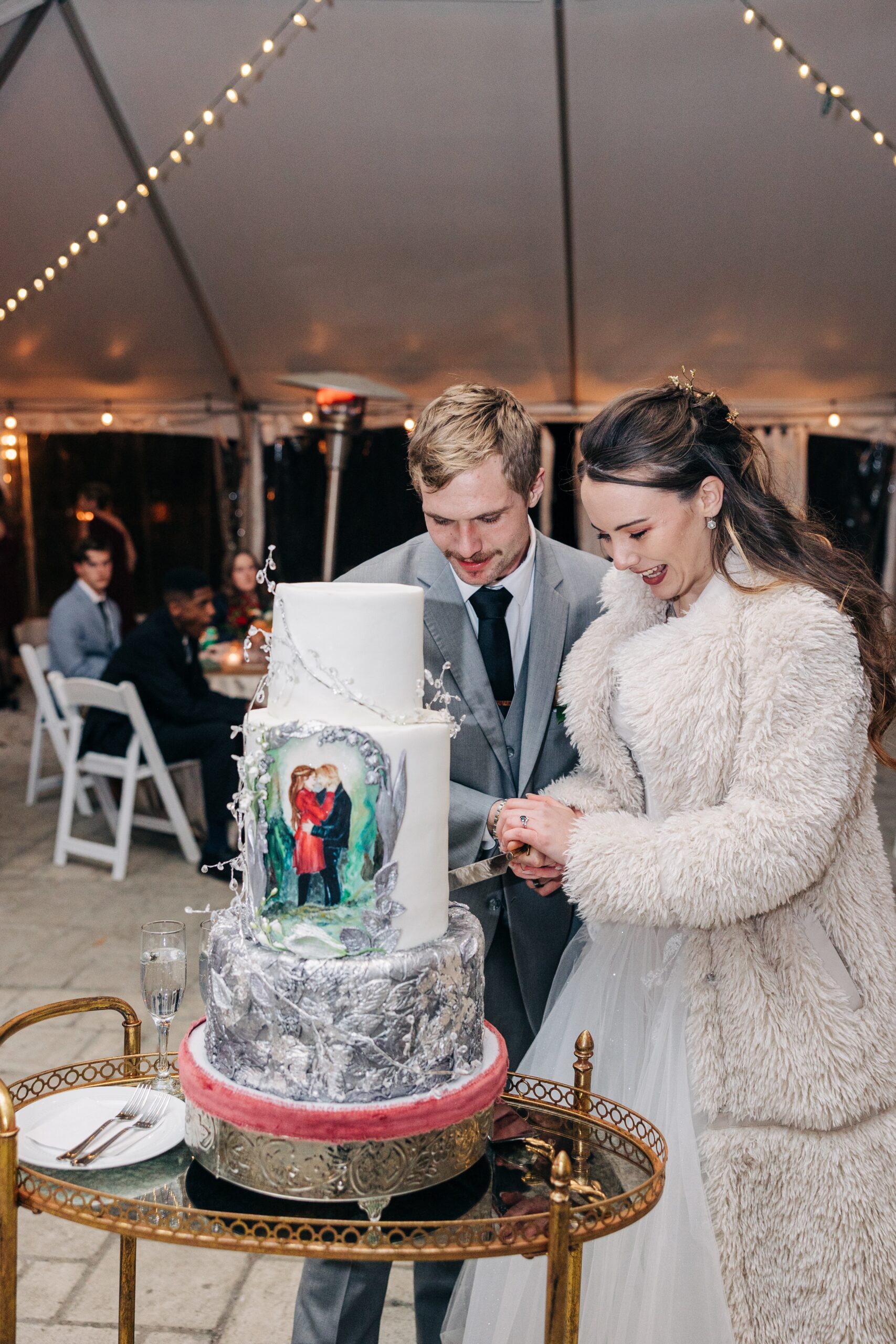 Newlyweds smile big while cutting their ornate decorated three tier cake under a tent