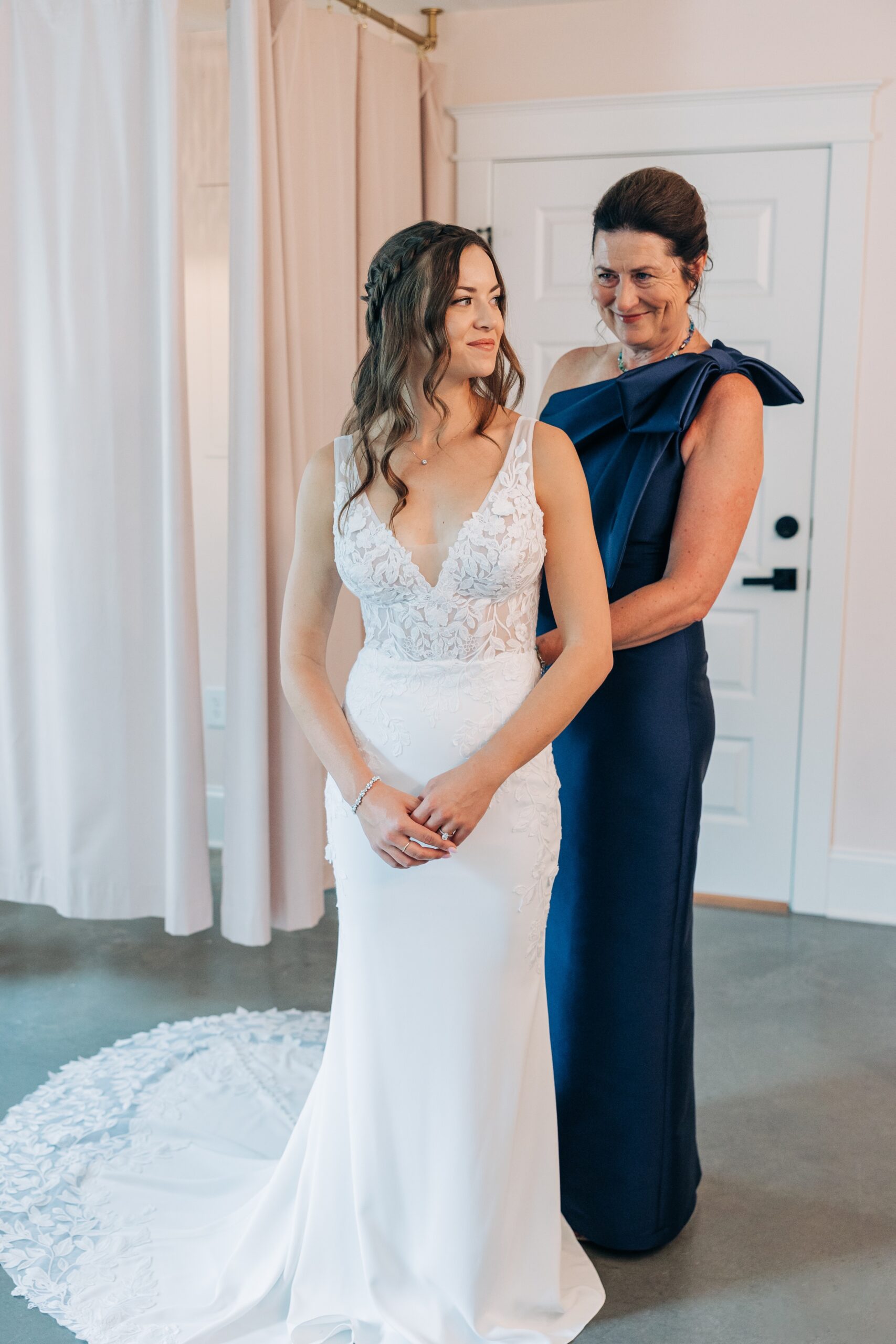 A bride smiles while mom helps button her into her lace dress