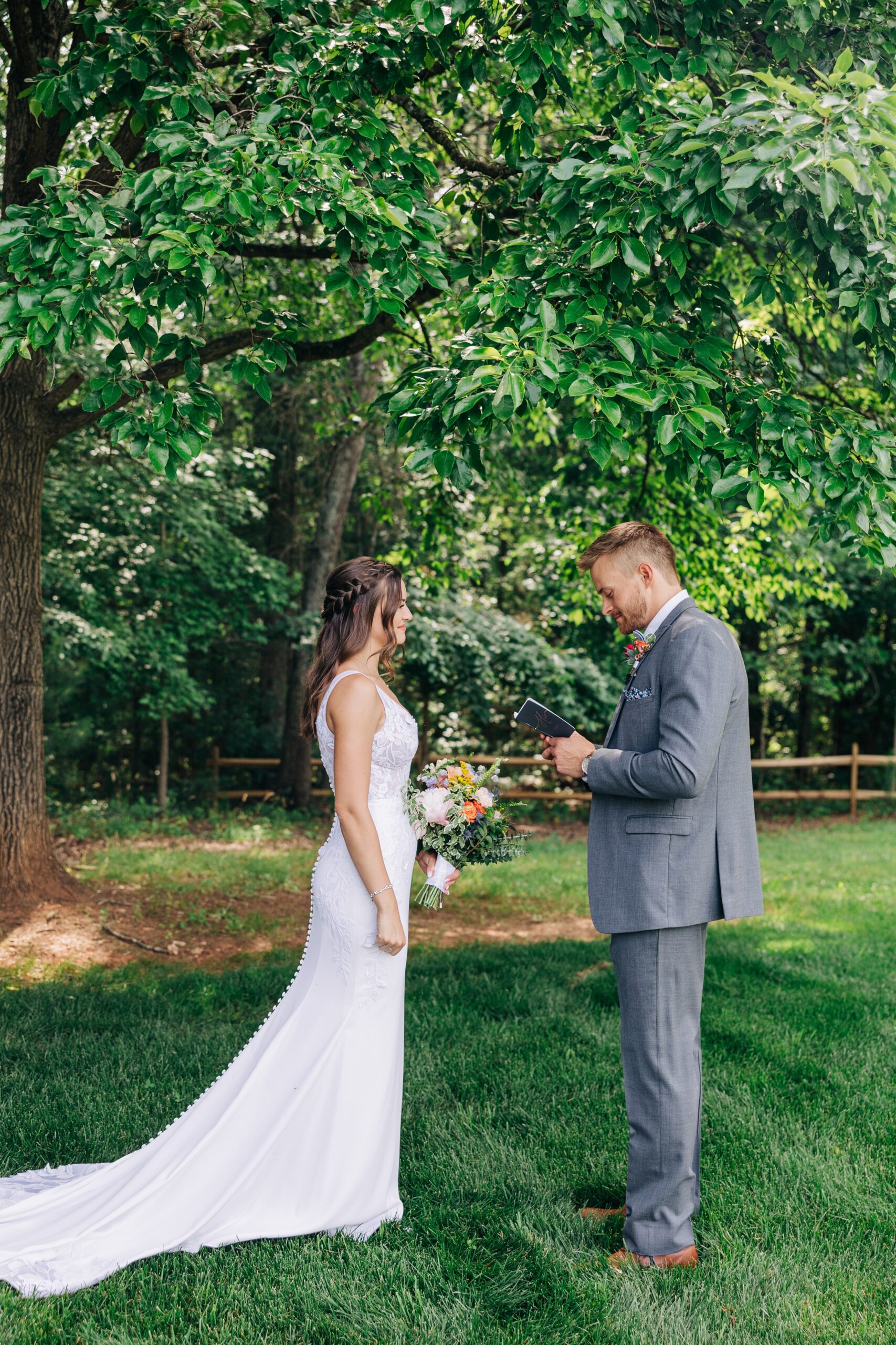 A groom reads his note to his bride under a tree in a lush lawn at The Ivory Barn