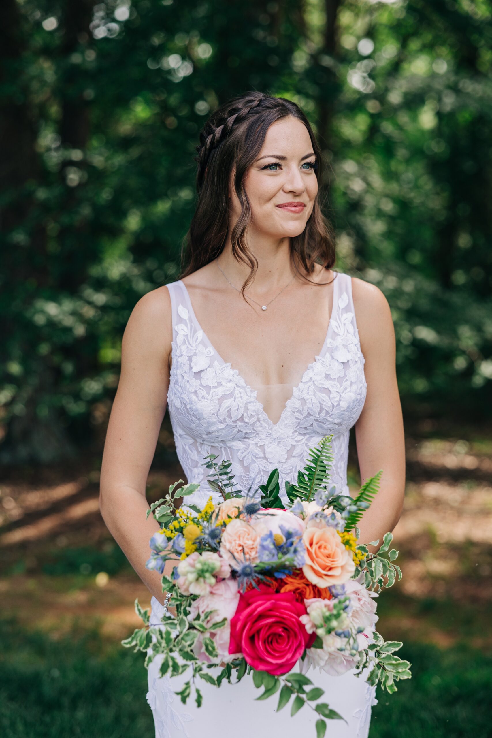 A happy bride stands in her lace dress holding her colorful bouquet on the edge of the forest