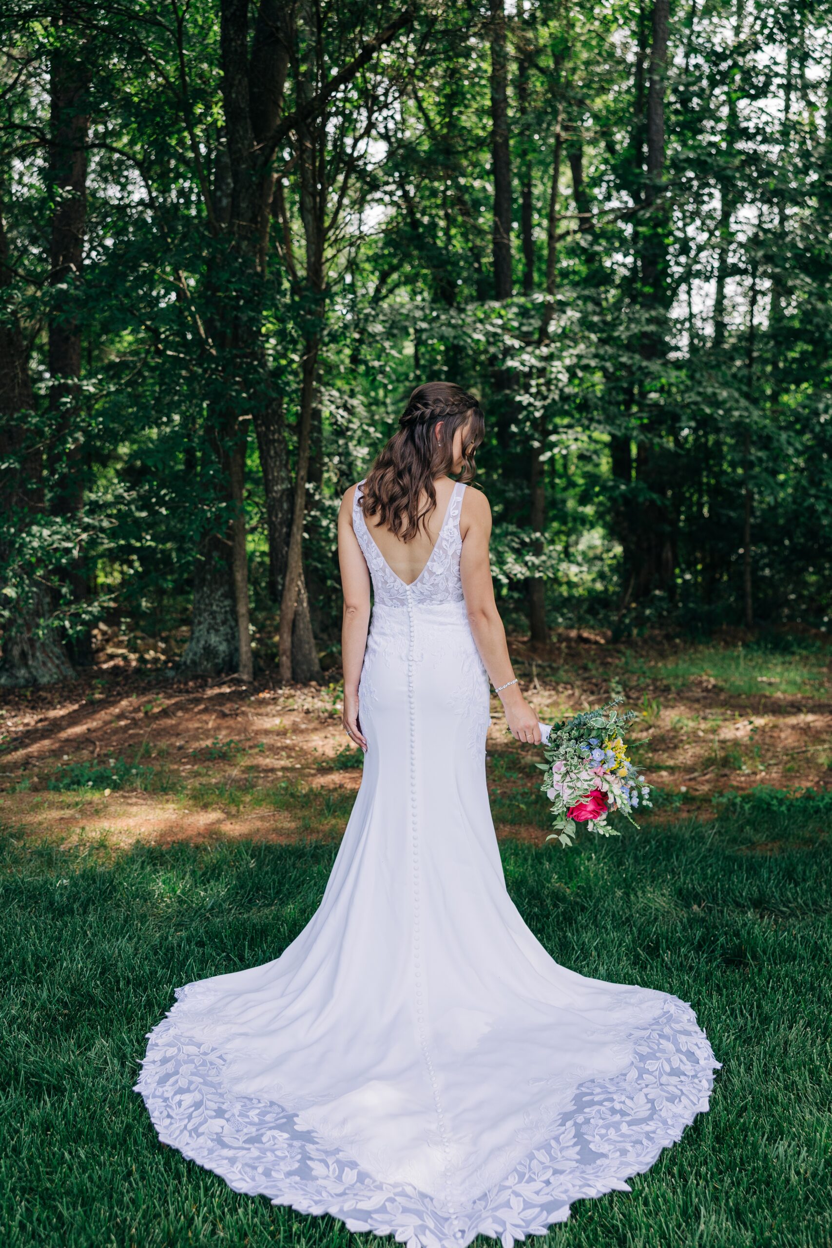 A bride shows off her lace train and colorful bouquet under some trees