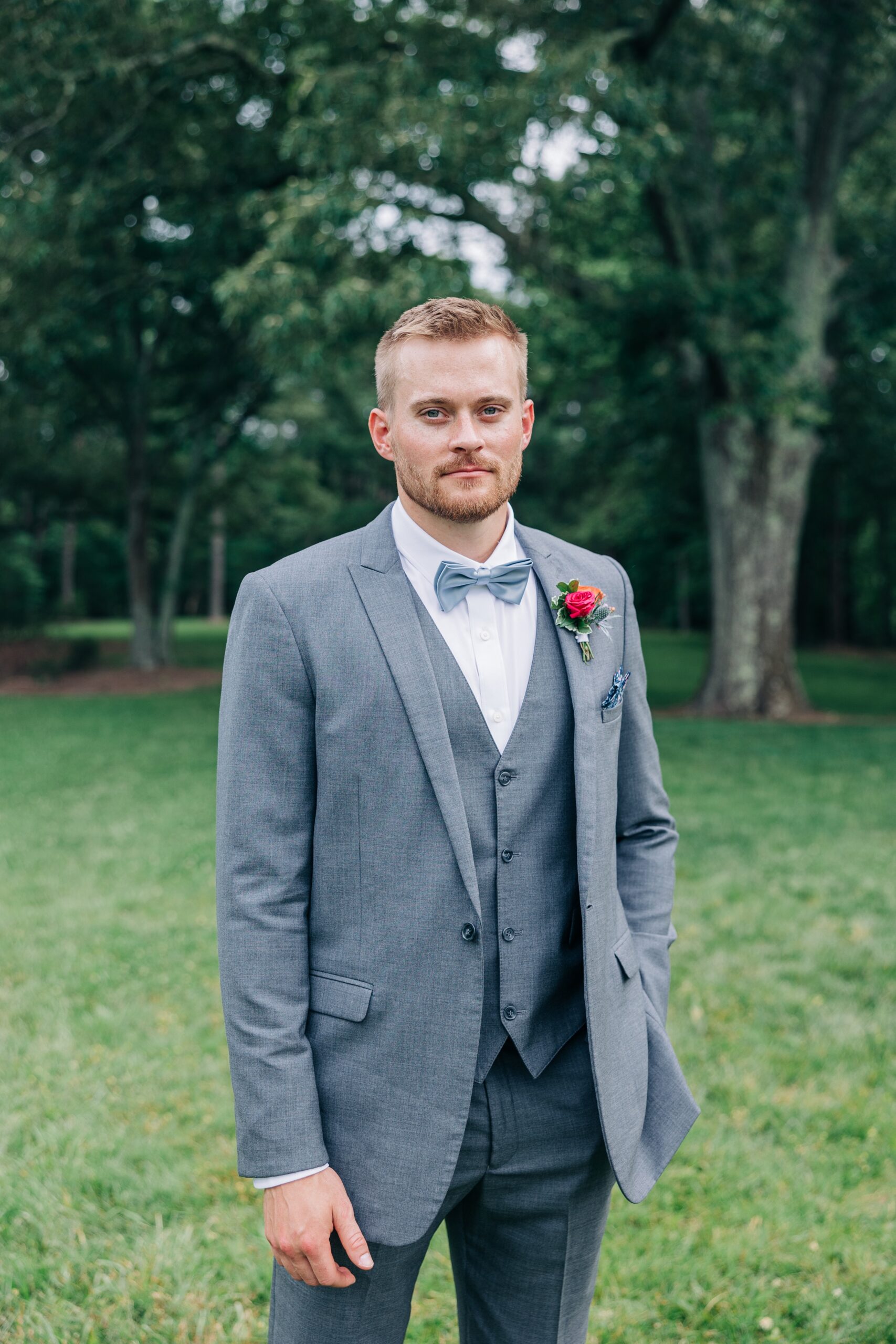 A groom stands in a grey suit with a red boutonnière and blue bowtie