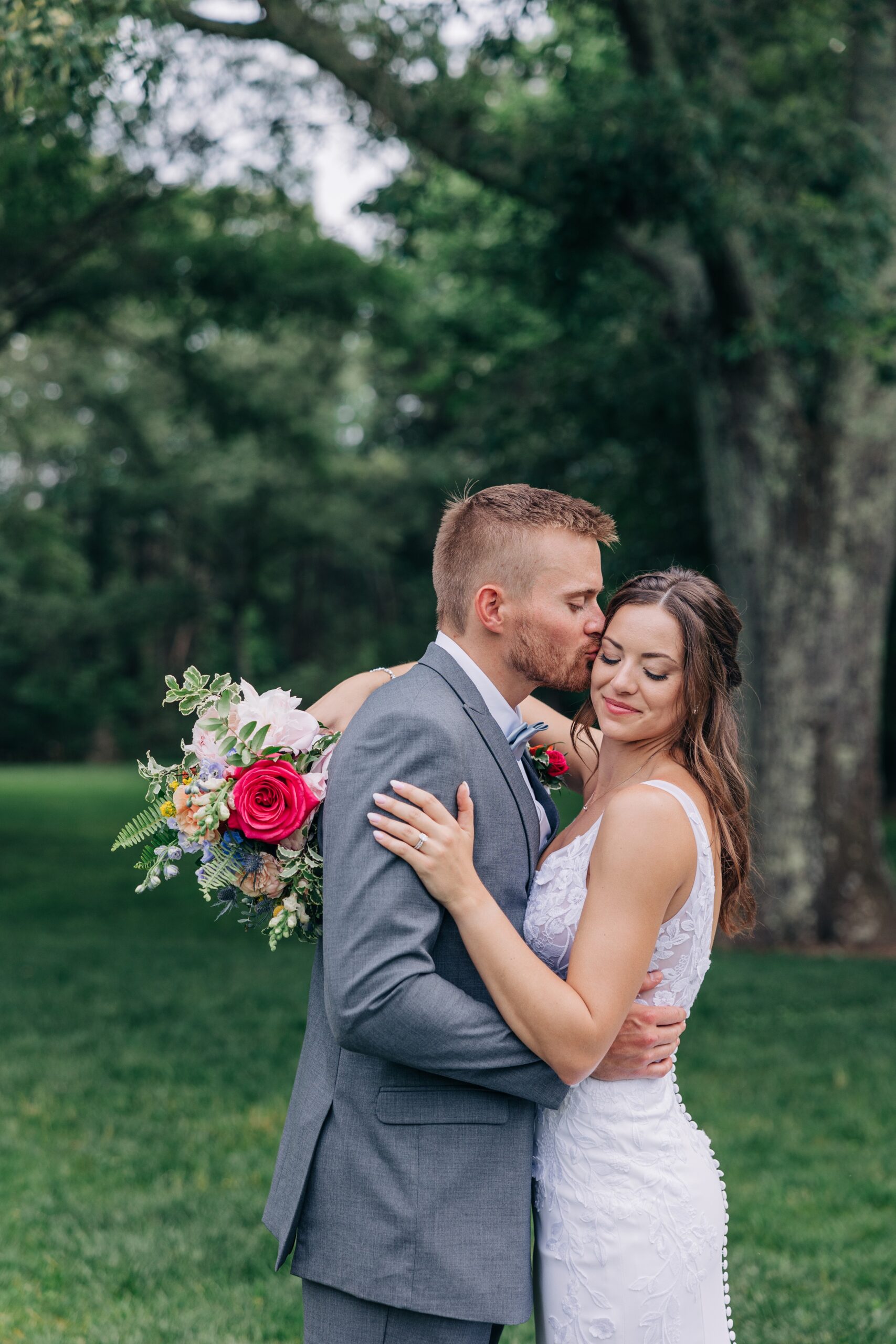 A groom kisses his bride's cheek as she hugs him and smiles down her shoulder