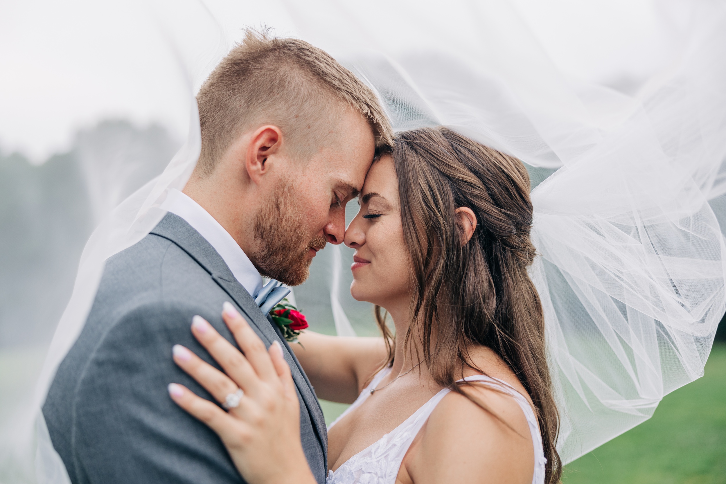 A happy bride and groom touch foreheads while snuggling under the long veil in the lawn