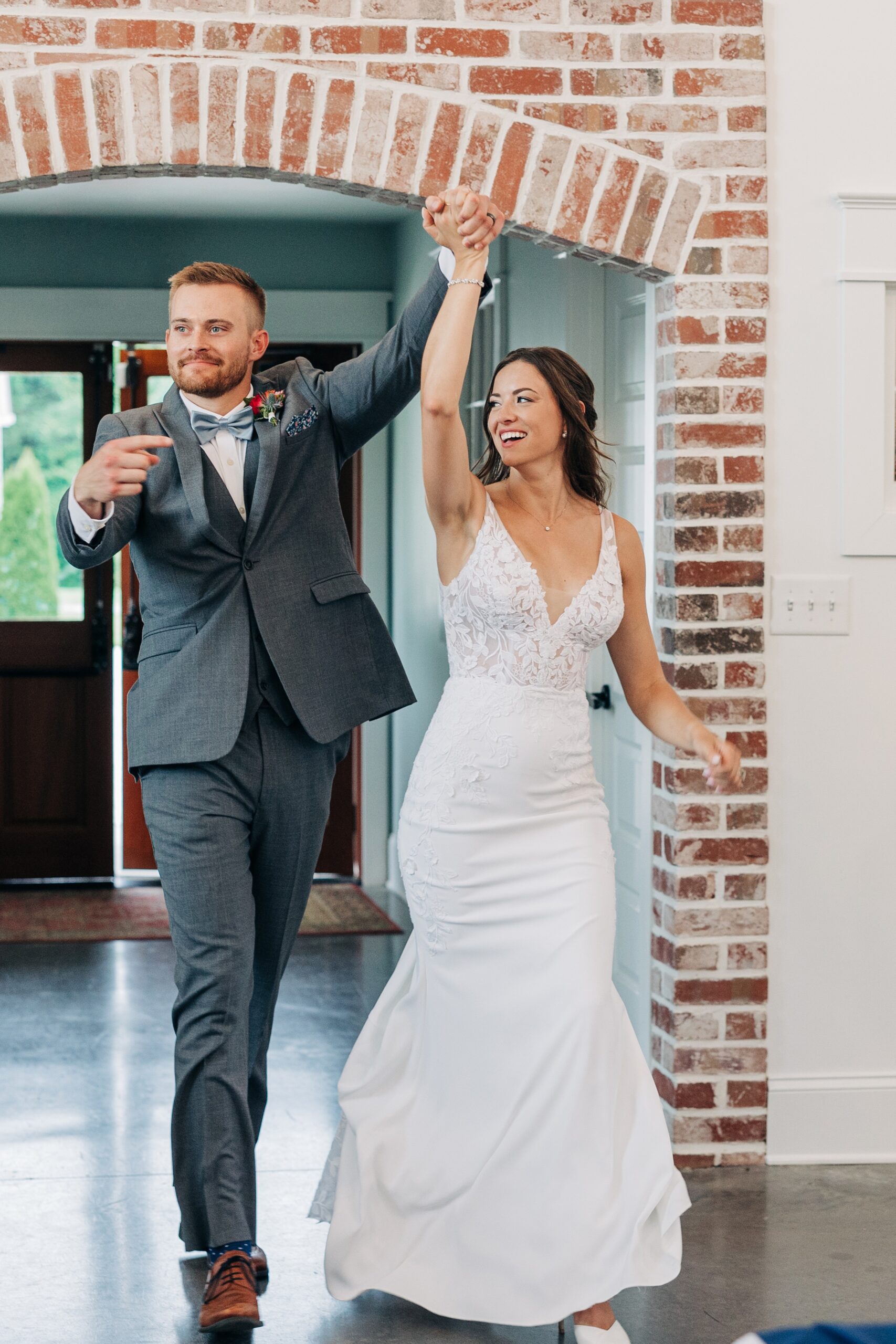 A groom in a grey suit lifts the hand of his bride as they enter their reception