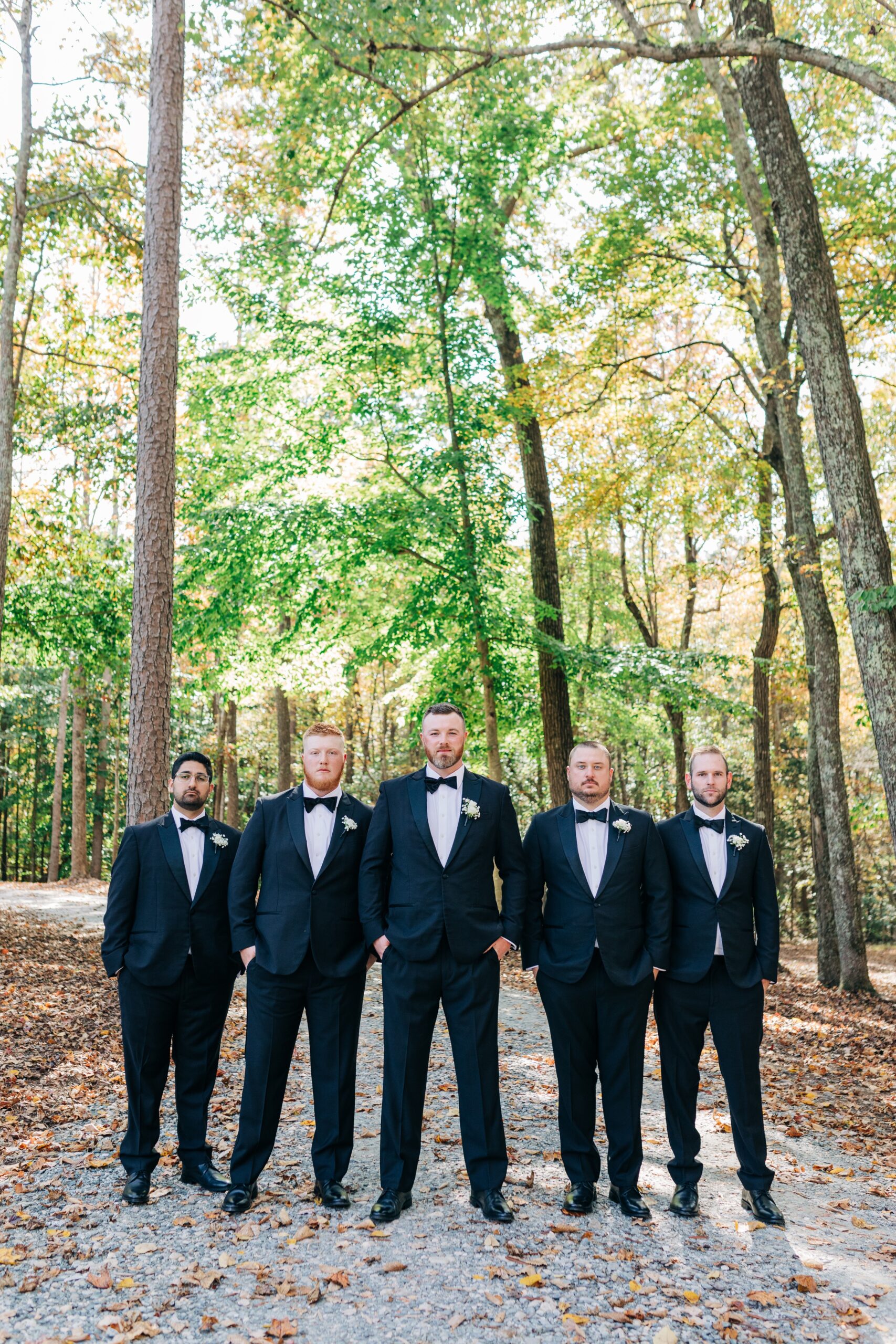 A groom stands in a gravel path in the forest with his groomsmen in black tuxes