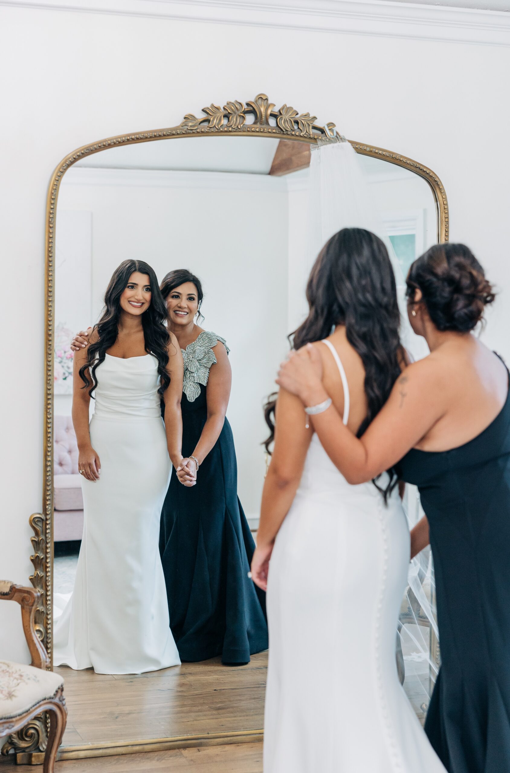 A bride stands in a mirror with a bridesmaid in her dress