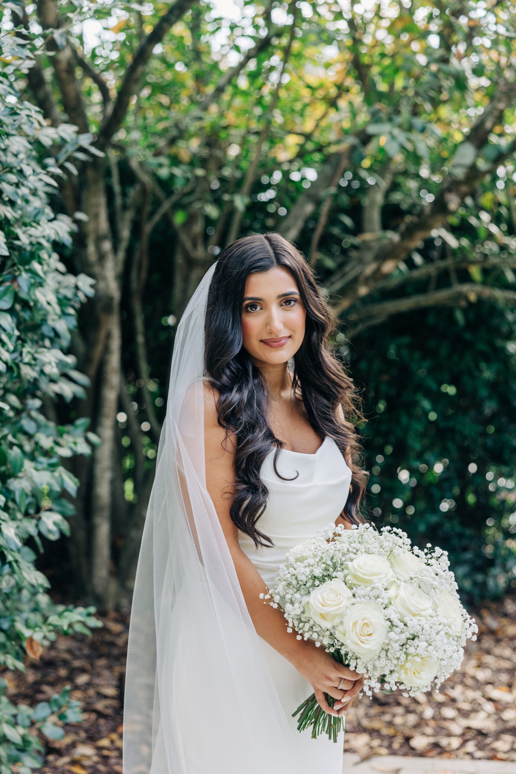 A bride smiles while standing in the gardens holding her white rose bouquet