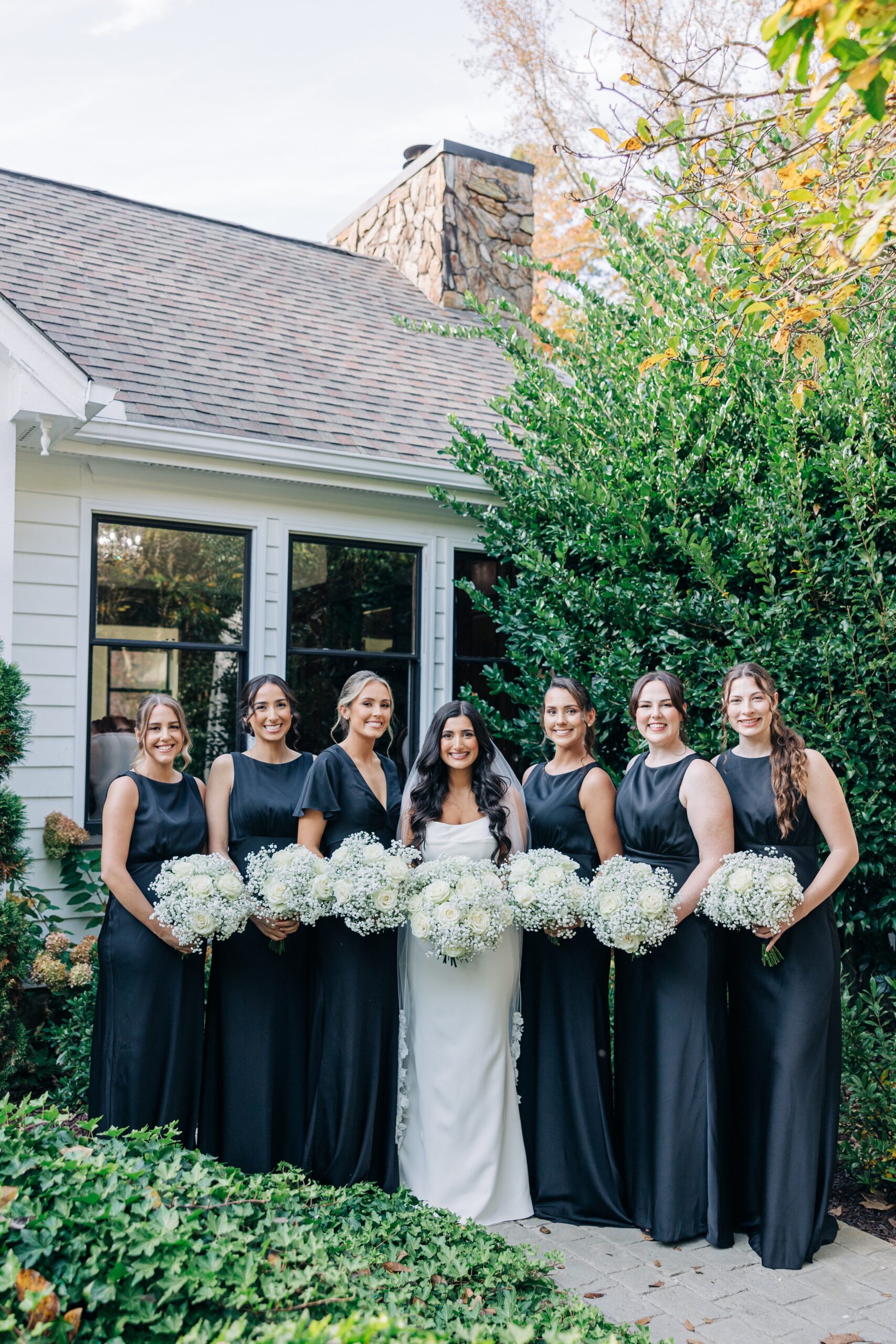 A bride smiles with her bridesmaids in black dresses hodling white bouquets