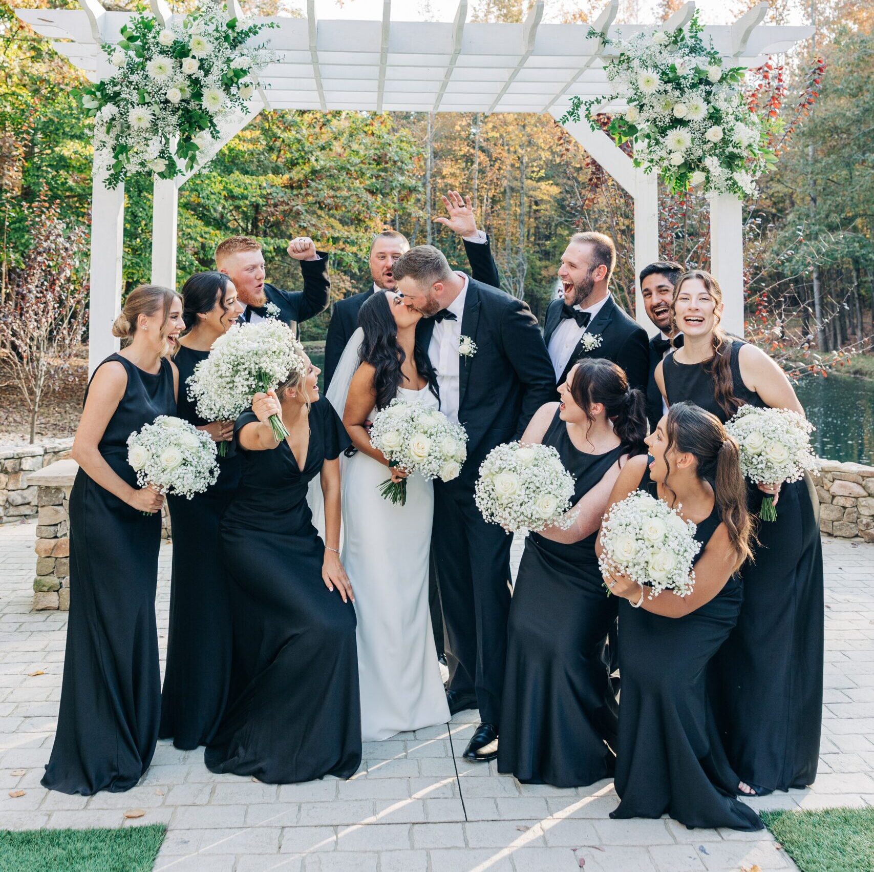 Newlyweds kiss under the pergola with their wedding party celebrating at their covington gardens wedding