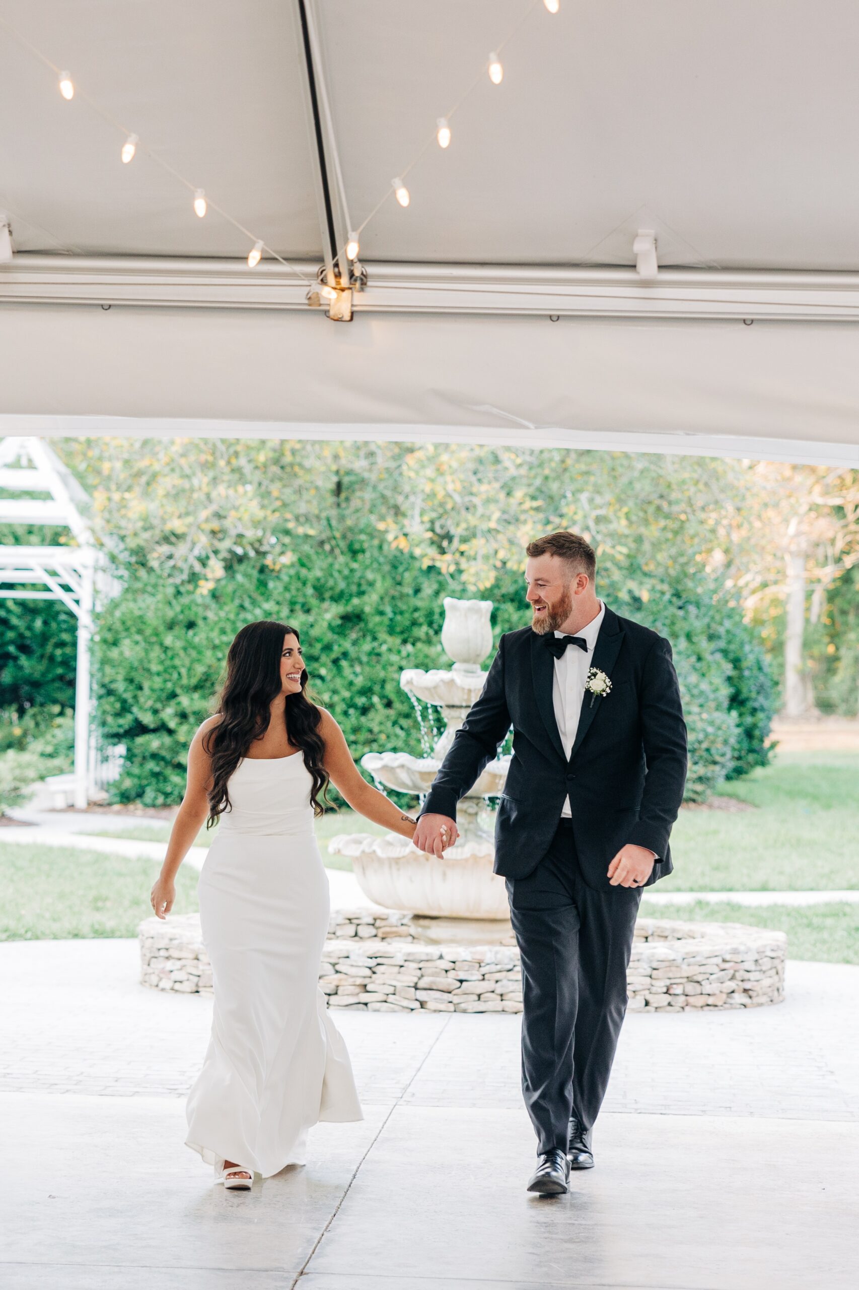 Newlyweds walk hand in hand to enter their outdoor covington gardens reception under a tent