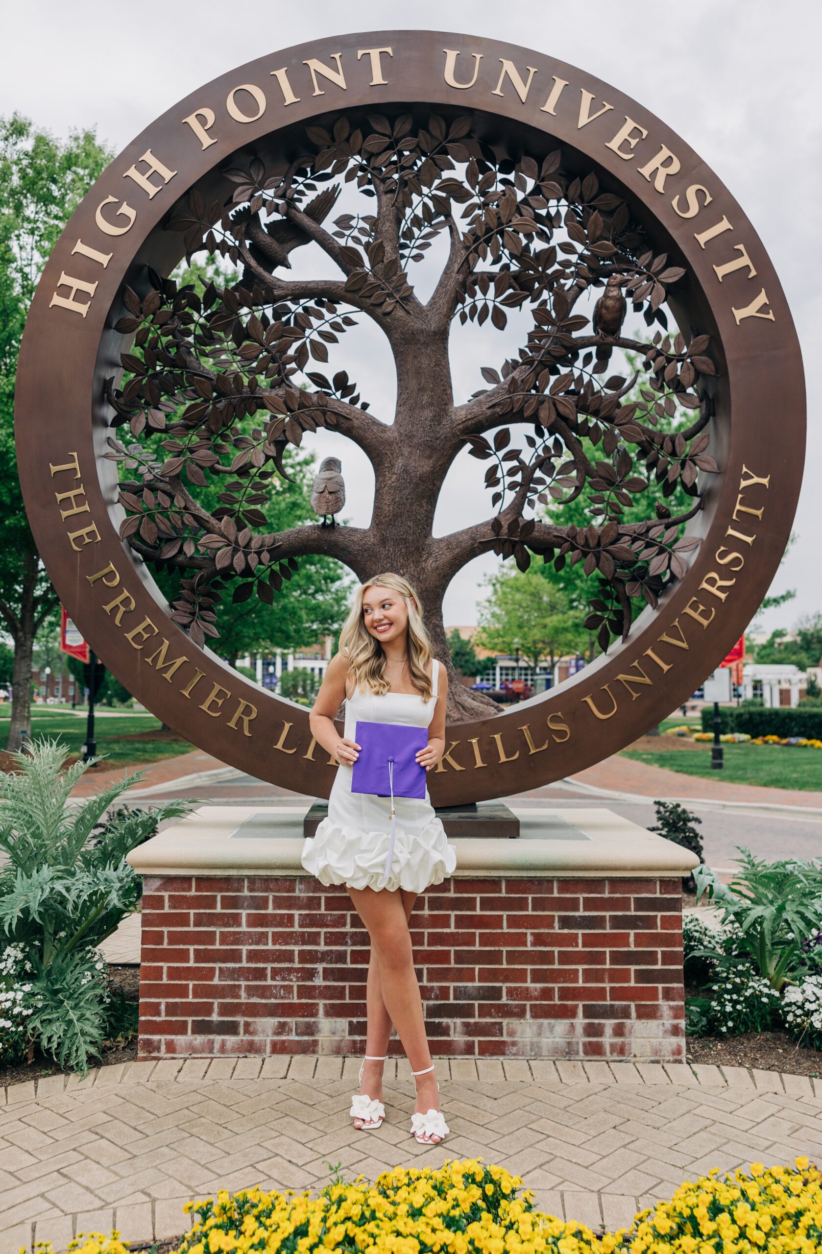 A happy grad holds her purple cap in front of the tree HPU statue