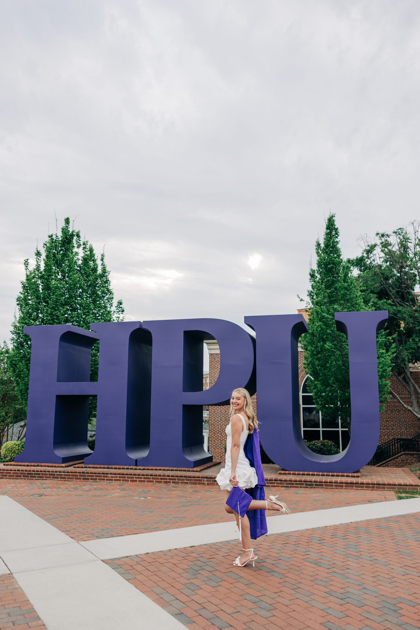 A graduate in a white dress pops her leg while walking in front of the HPU sign for her high point university graduation photographer