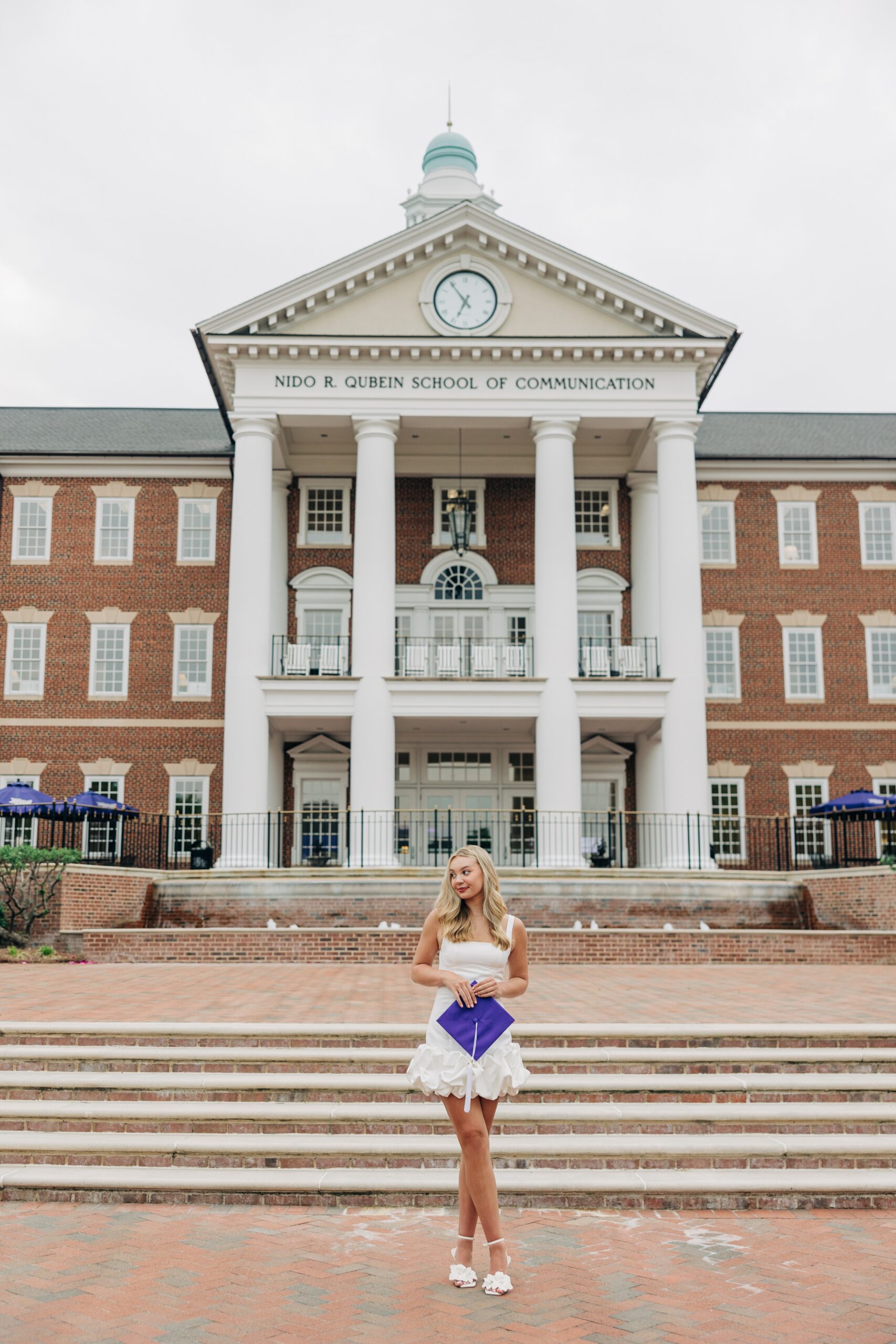 A woman in a white dress holds her purple cap while standing in front of the colonial communications building