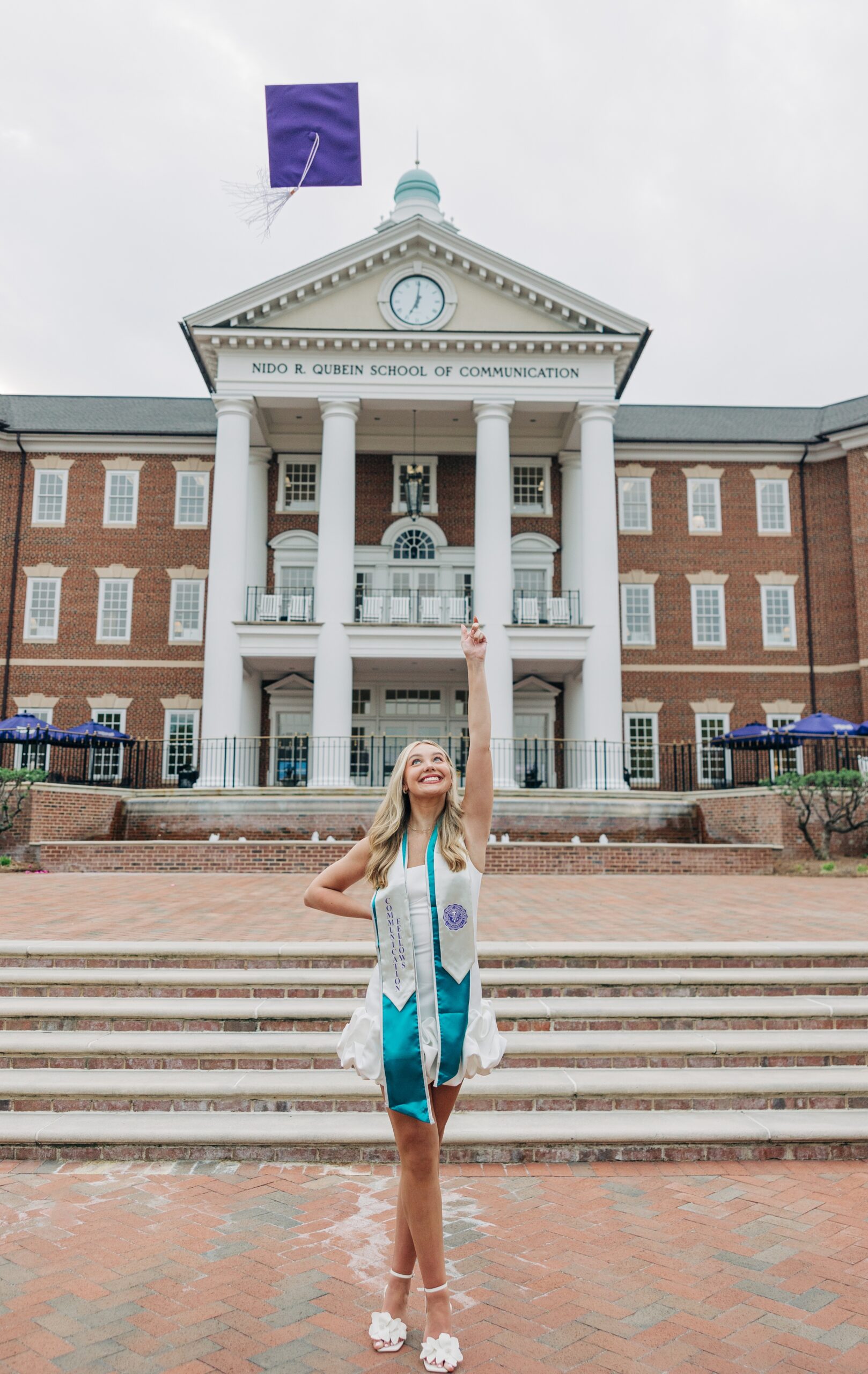 A grad tosses her purple cap in the air in front of the communication building in a white dress