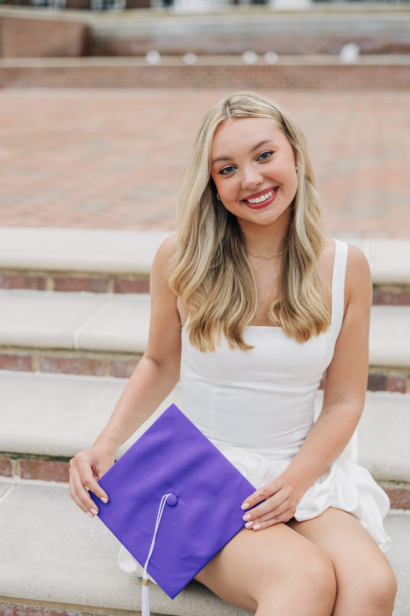 A smiling graduate in a white dress sits on brick steps by a fountain holding her purple cap