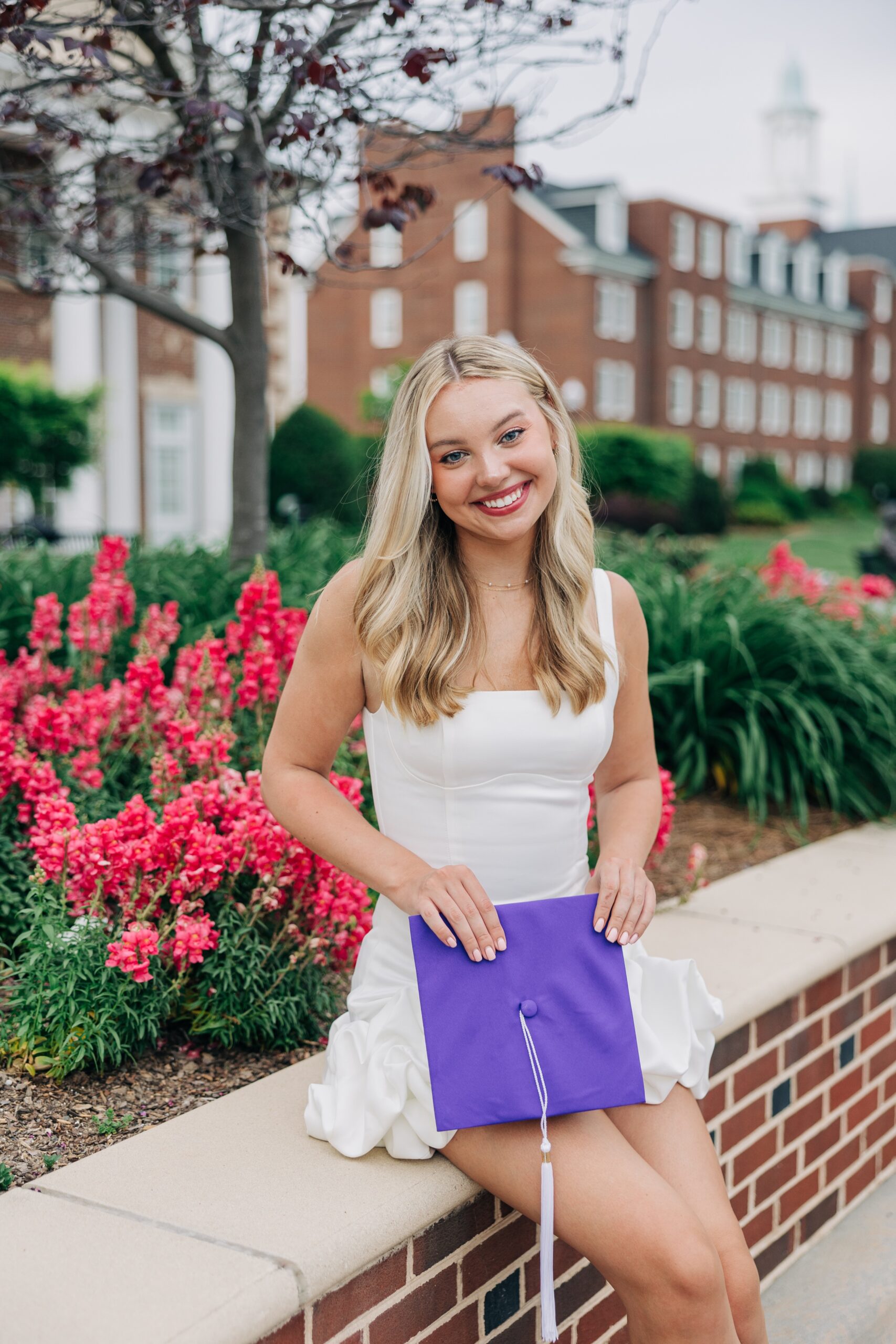 A happy blonde in a white dress smiles while sitting on a garden wall for her high point university graduation photographer