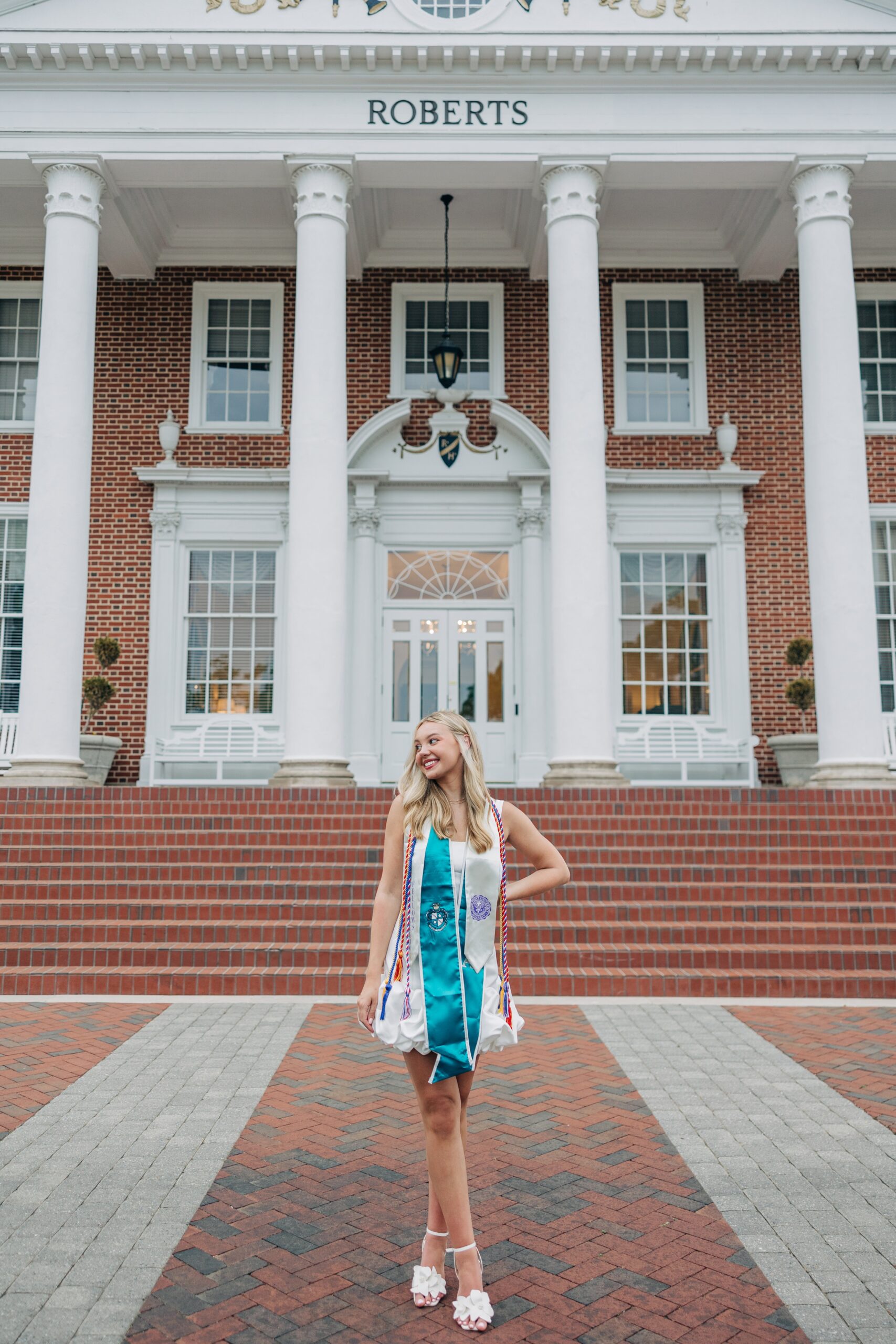 A happy grad struts on brick sidewalk in her sorority stole and white dress